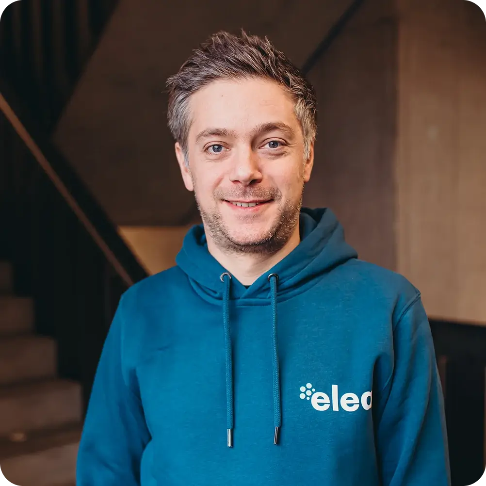 Lächelnder Mann mit kurz geschnittenem grauem Haar trägt einen blauen Kapuzenpullover mit Teil des Schriftzugs 'elec'.