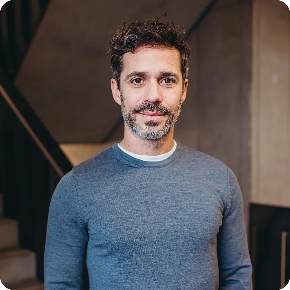 Mittelalter Mann mit lockigem braunem Haar und Bart, der grau-blauen Pullover trägt und in einem Innenraum steht.