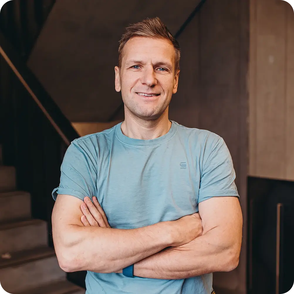 Mann in blauem T-Shirt steht mit verschränkten Armen vor einer Treppe und lächelt.