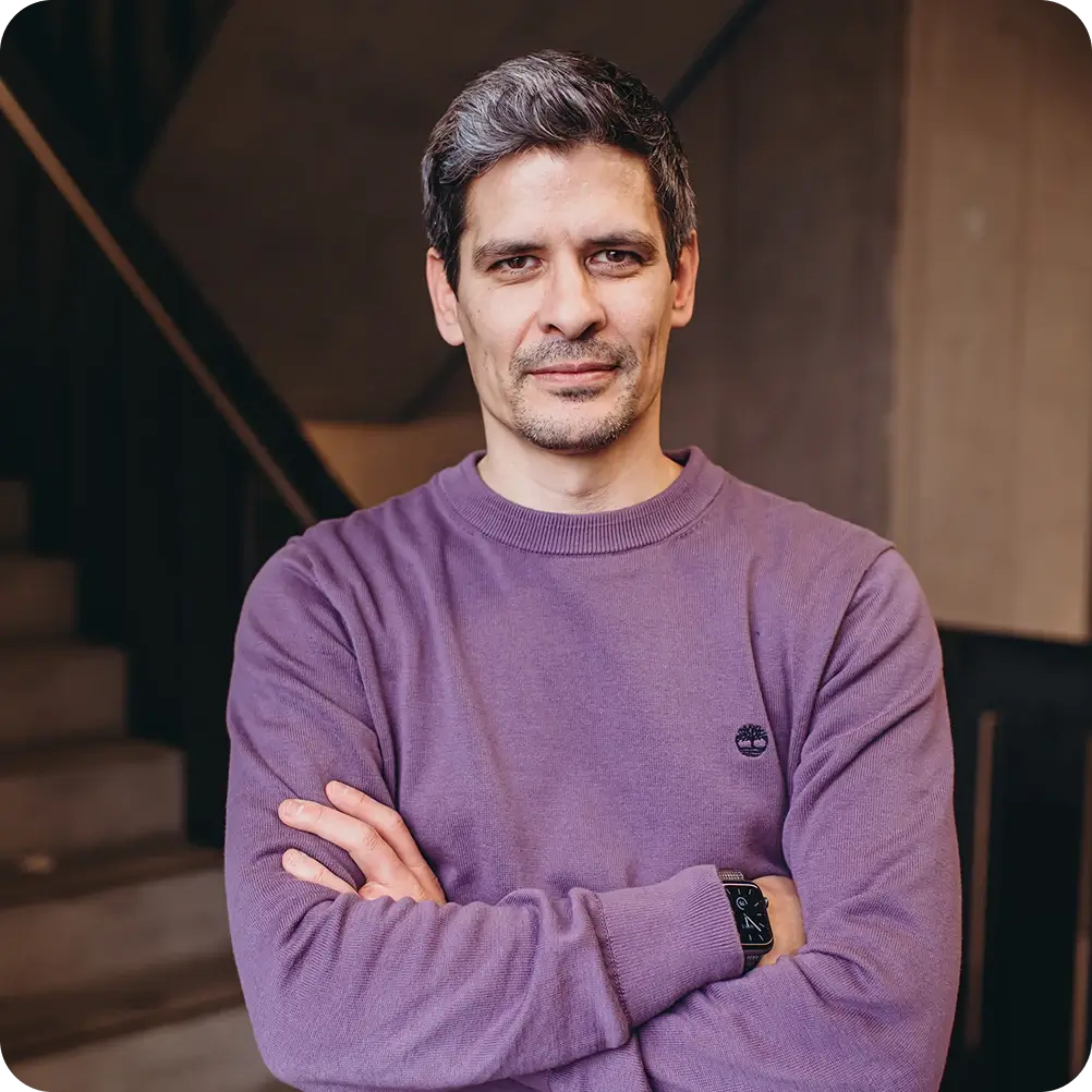 Mann mit graumeliertem Haar und lila Pullover steht mit verschränkten Armen vor einer Treppe in Innenraum.