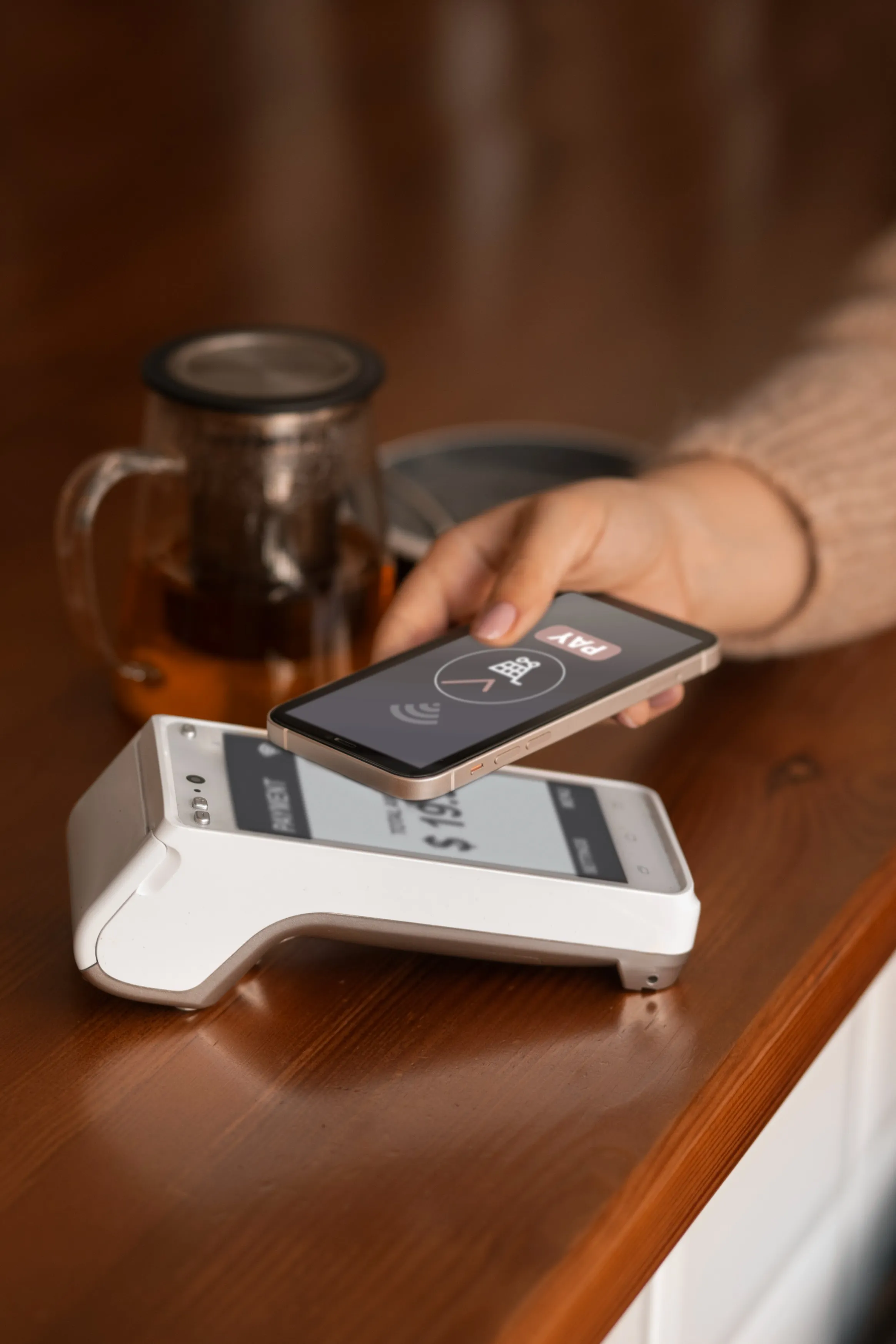 PayTrac | Person holding a smartphone showing a 'PAY' screen near a card payment terminal on a wooden table.