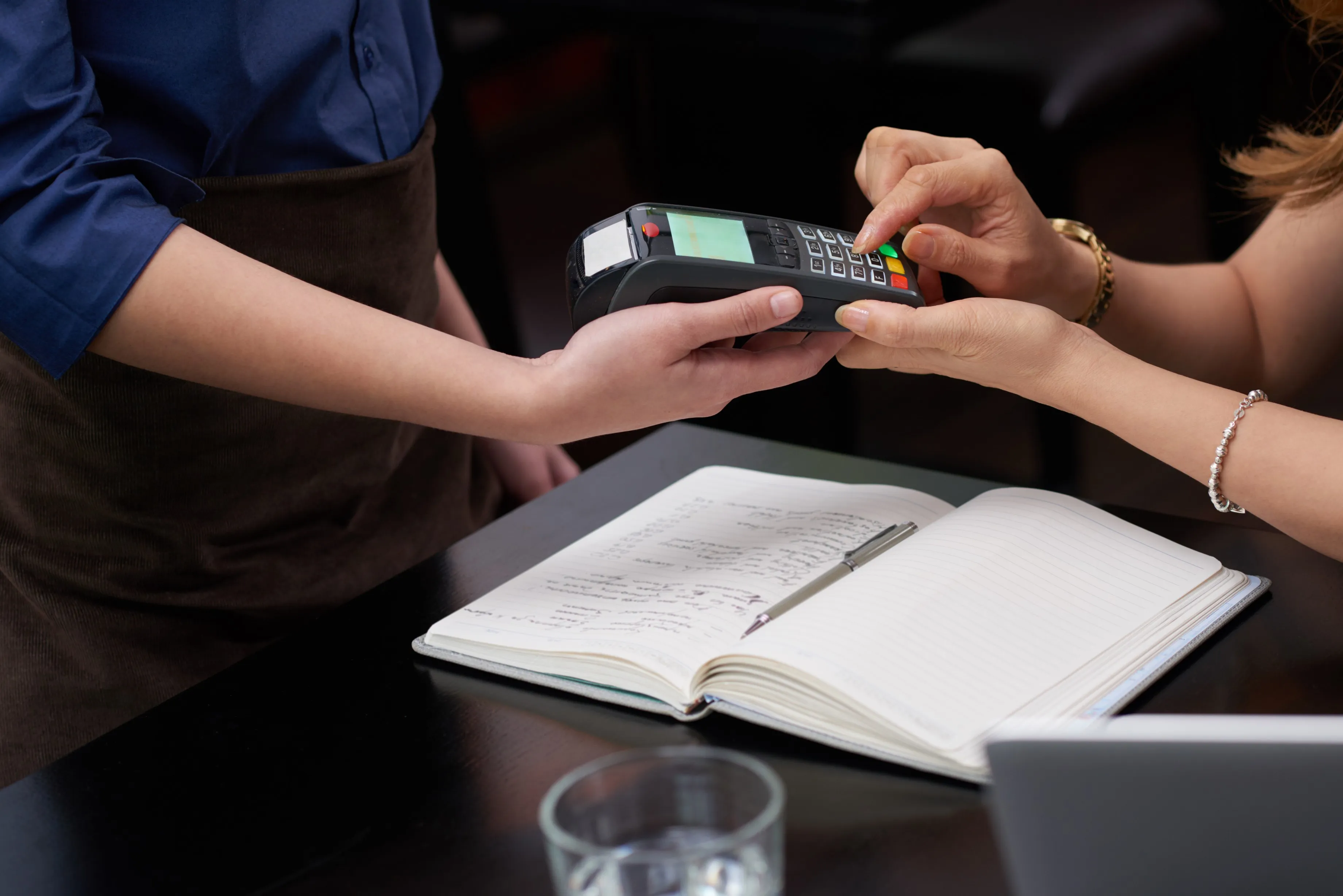PayTrac | Customer entering PIN on a payment terminal held by a person wearing a brown apron, with an open notebook and pen on a black table nearby.