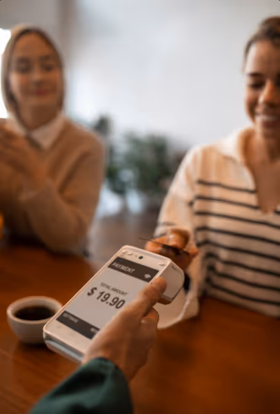 Person holding a payment terminal showing a total amount of $19.90, while another person prepares to tap a credit card for payment.
