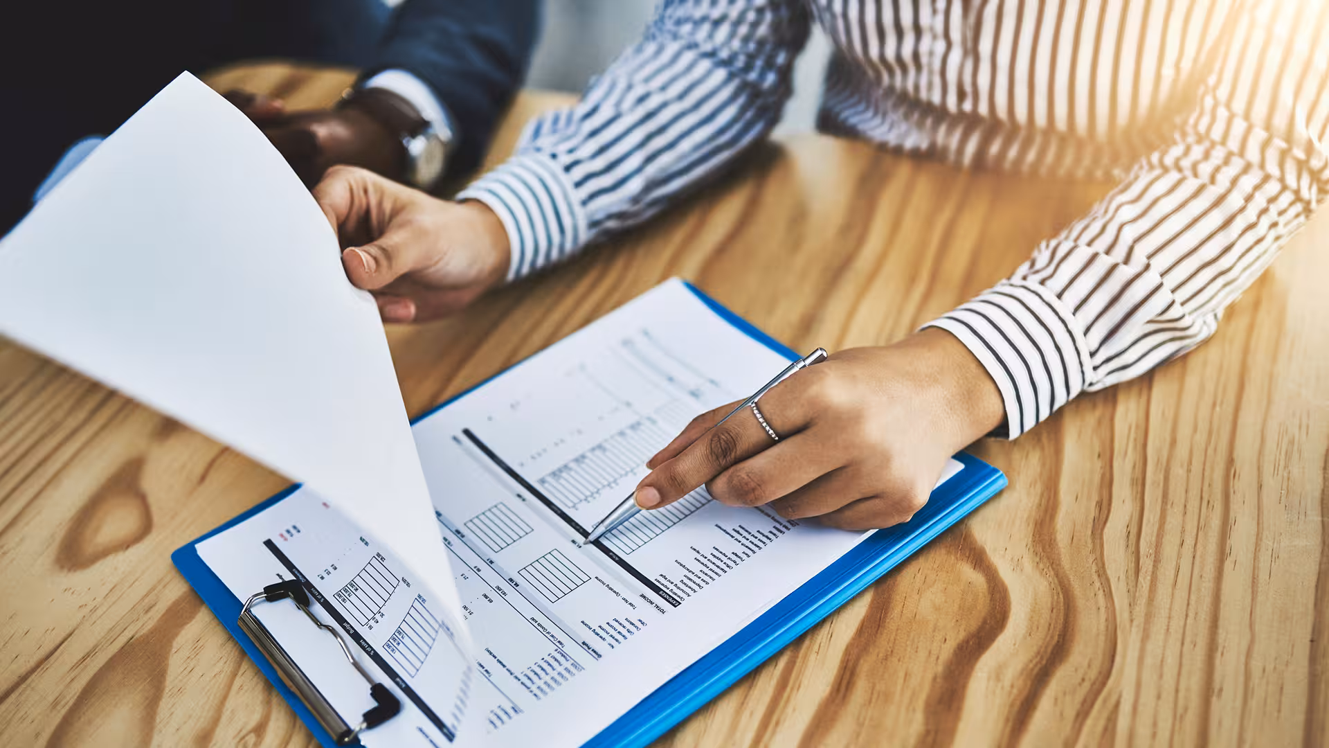 Hands consulting a report on a wooden desk with other documents