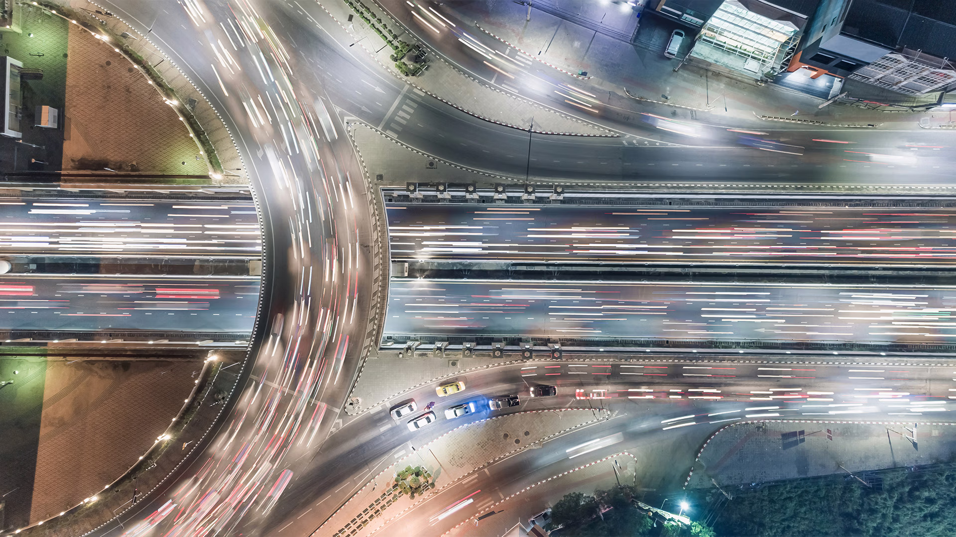 Image visible : vue aérienne en plongée (vue du dessus), de nuit, d'un grand échangeur/carrefour routier — chaussées courbes et voies parallèles parcourues de traînées lumineuses (pose longue) matérialisant le flux des véhicules, quelques voitures nettes à l'arrêt, bâtiments et végétation en périphérie.