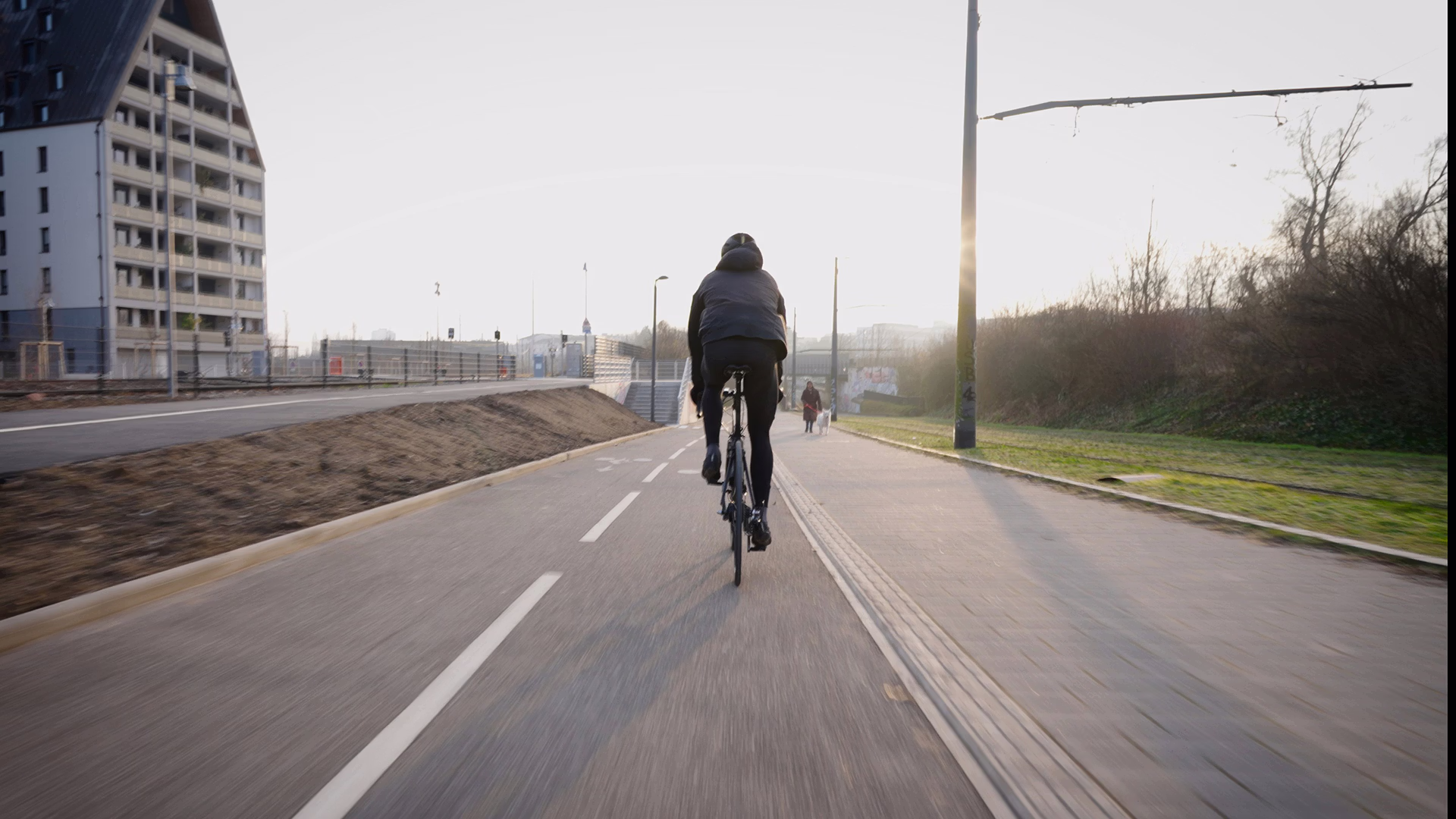 Cycliste vu de dos roulant sur une piste cyclable urbaine au lever du soleil, longeant un immeuble et une voie verte