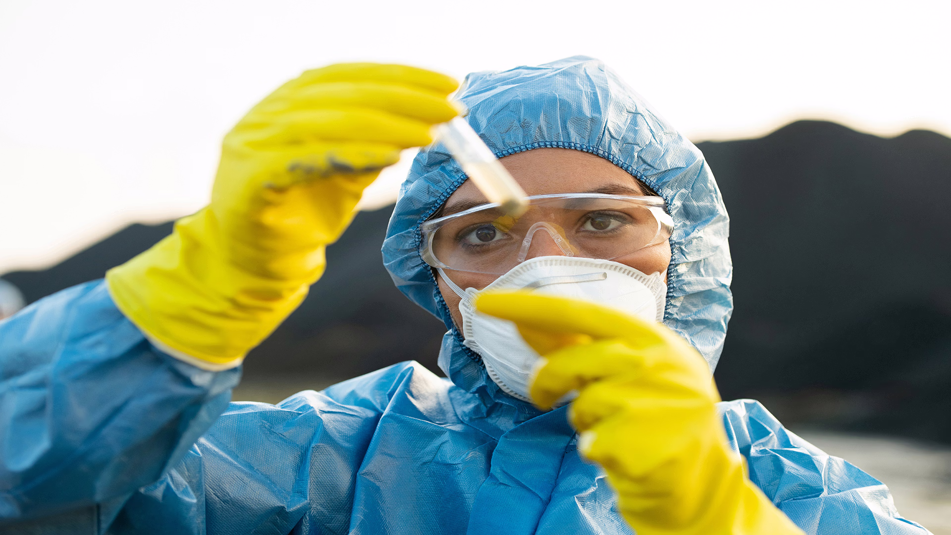 Technicienne en combinaison de protection, masque et gants jaunes examinant un échantillon dans une éprouvette sur le terrain