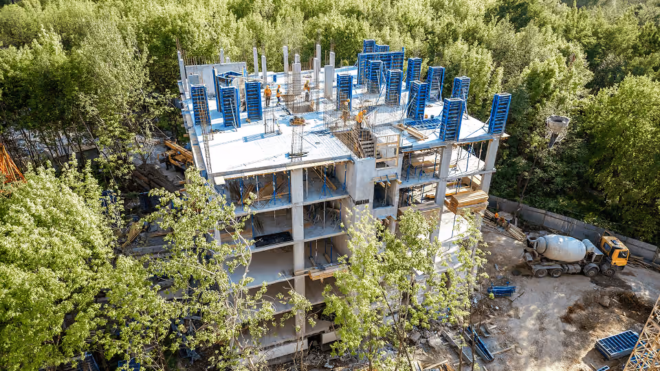 Chantier de construction d'un immeuble en béton entouré d'arbres verts, avec bétonnière sur site.