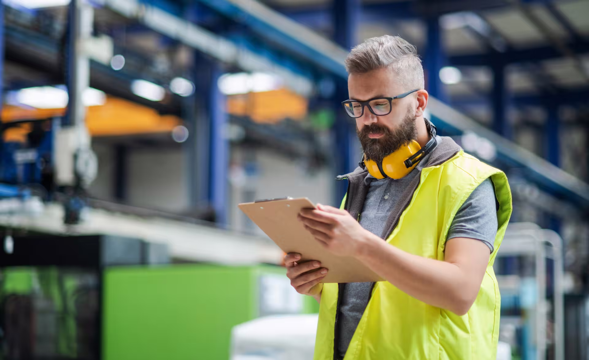 Technicien barbu à lunettes, portant un gilet de sécurité jaune fluo et un casque anti-bruit jaune autour du cou, consultant un porte-bloc dans un environnement industriel dont l'arrière-plan flou laisse apparaître des structures métalliques.