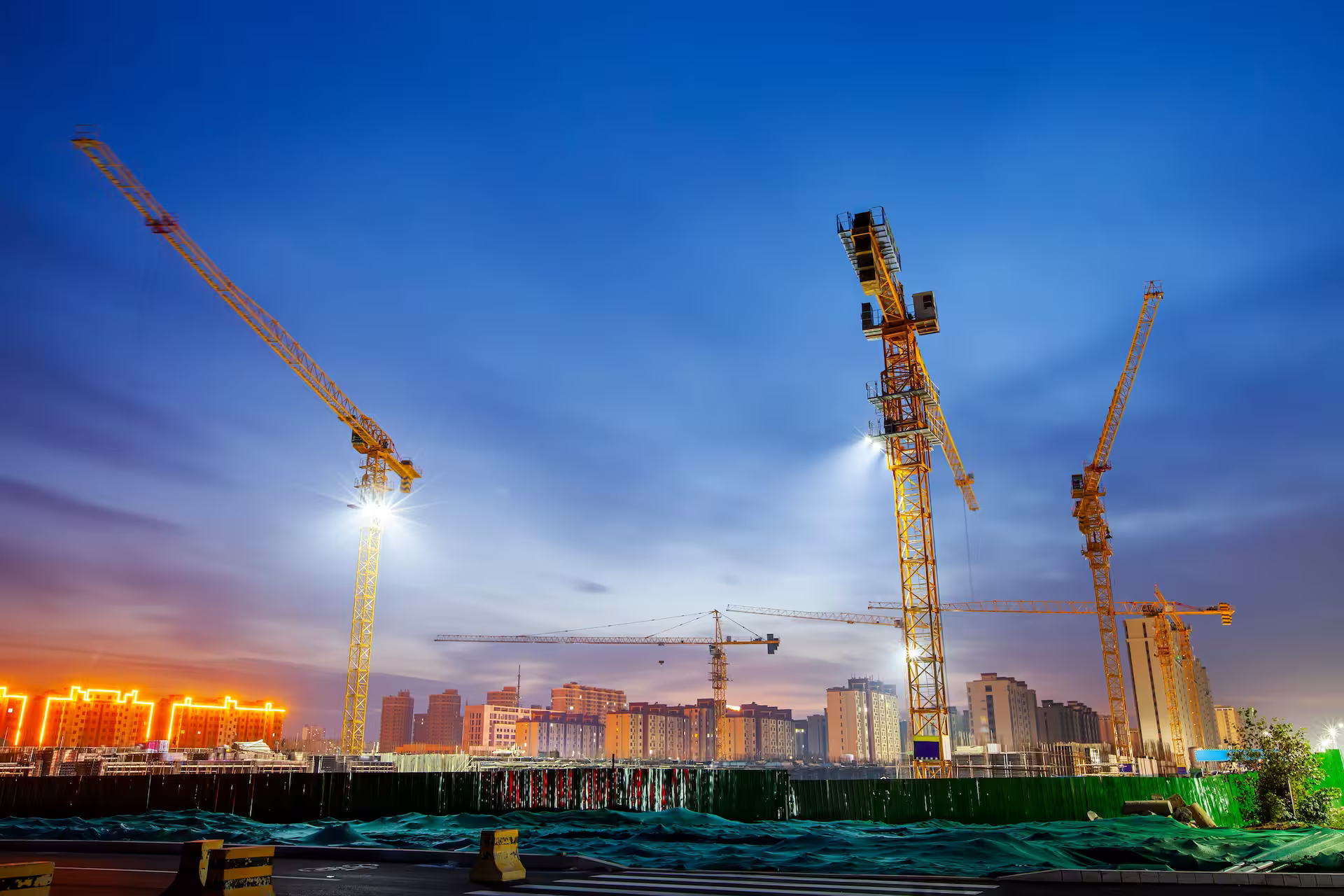 Several illuminated yellow tower cranes on an urban construction site at dusk, with buildings in the background under a blue and orange sky.