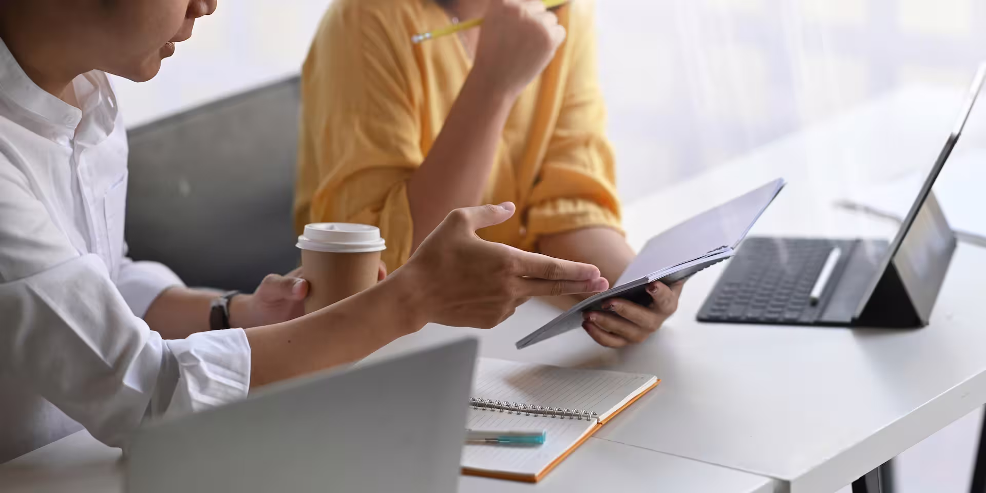 Deux collègues échangent autour d'un document lors d'une réunion de travail, avec ordinateurs portables, carnet et café sur la table.