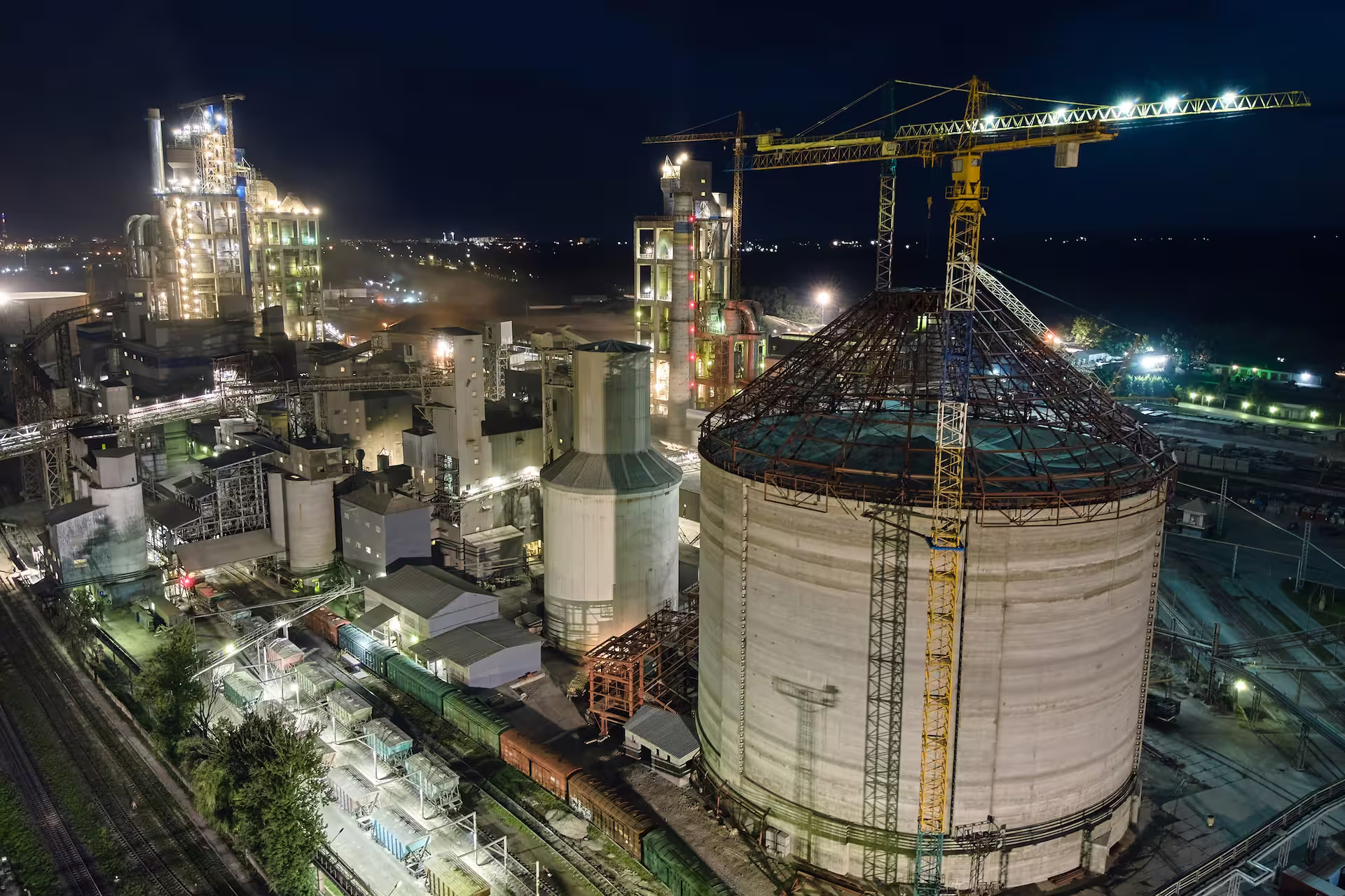 Vue aérienne nocturne d'une cimenterie éclairée, avec tours de production, silos en construction, grues et wagons de fret.
