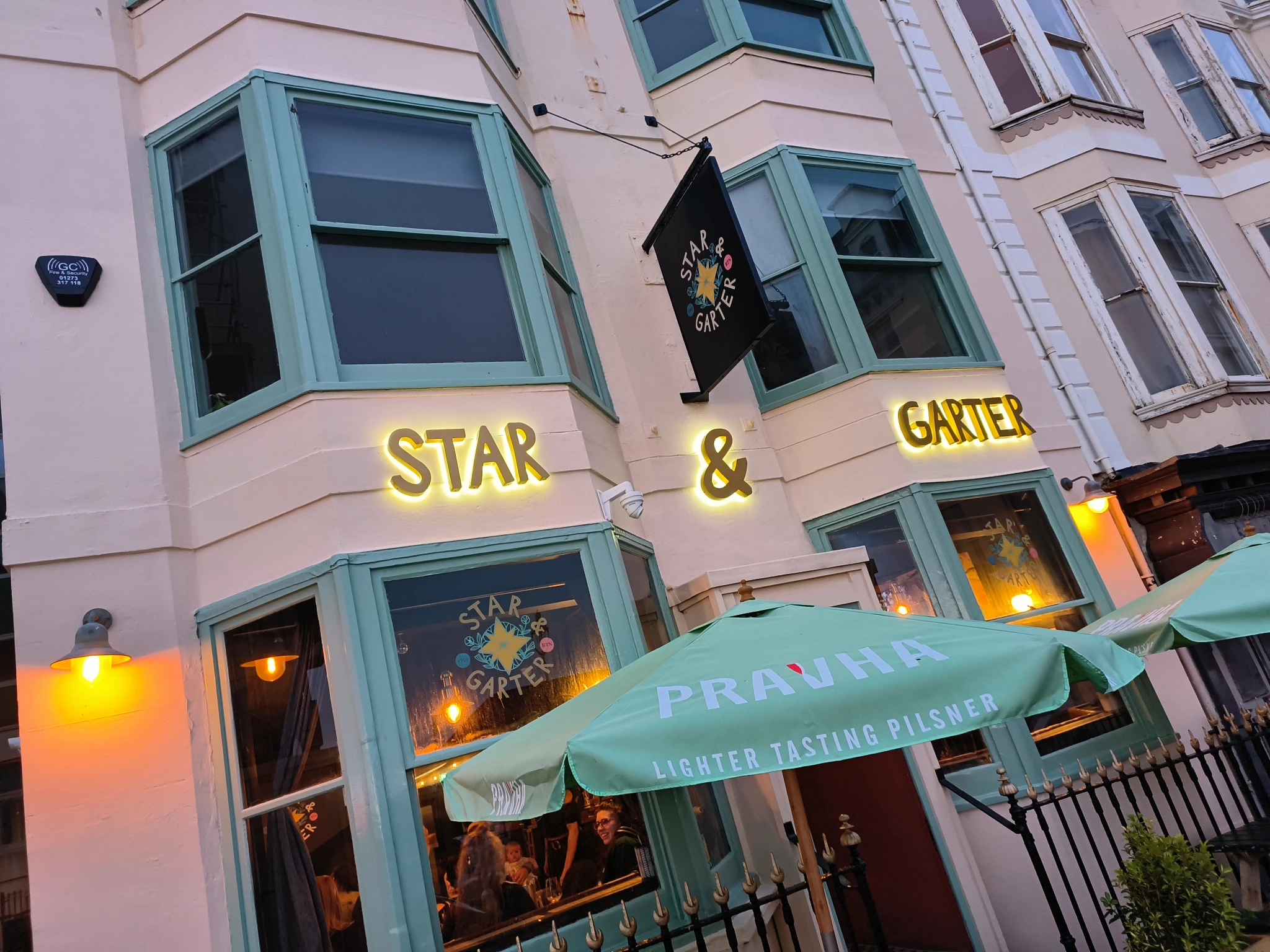 Exterior of Star & Garter pub with lit yellow sign and green umbrellas reading Praha lighter tasting pilsner.