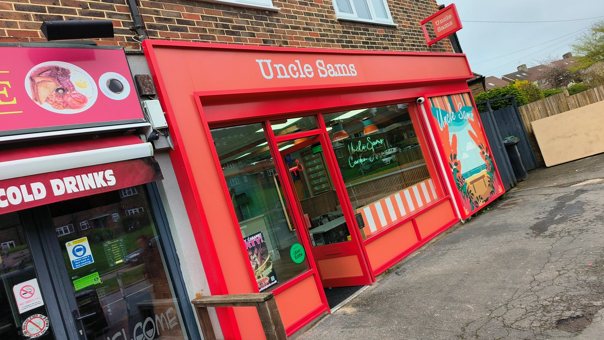 Bright red storefront of Uncle Sams with glass door, window art, and adjacent shop advertising cold drinks.