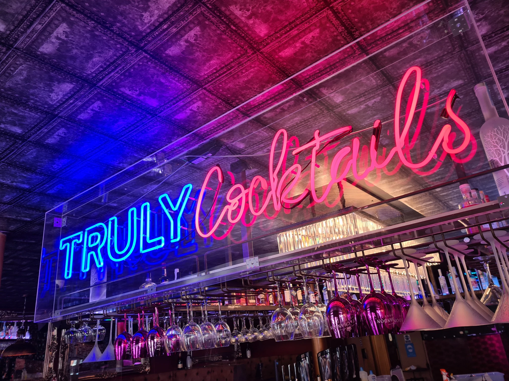 Neon sign reading 'TRULY Cocktails' above hanging wine glasses in a bar with a dark ornate ceiling.