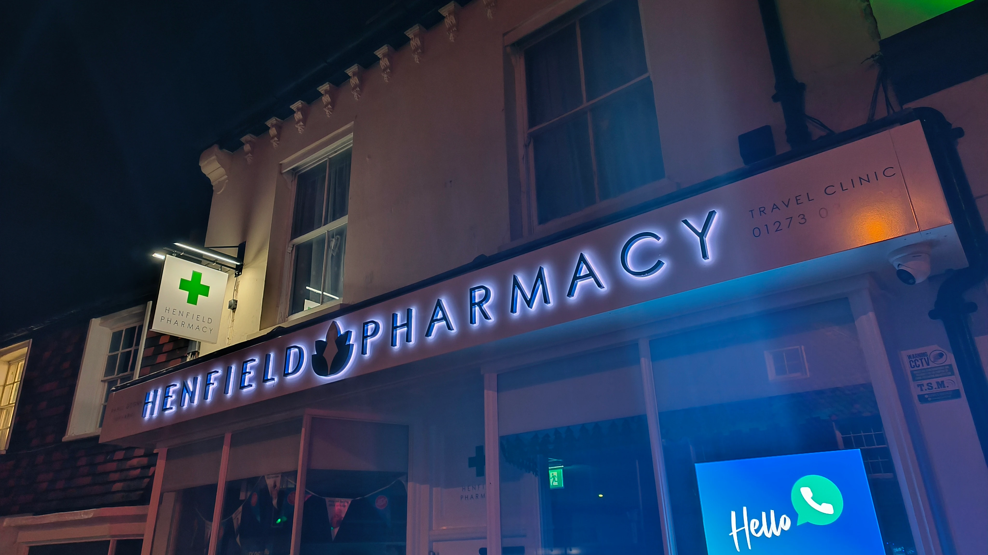 Henfield Pharmacy store sign lit up at night with a green cross symbol and a lit Hello phone icon.