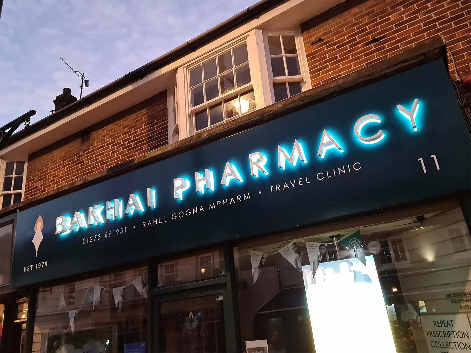 Exterior view of Bakhai Pharmacy with illuminated sign and brick building facade at dusk.