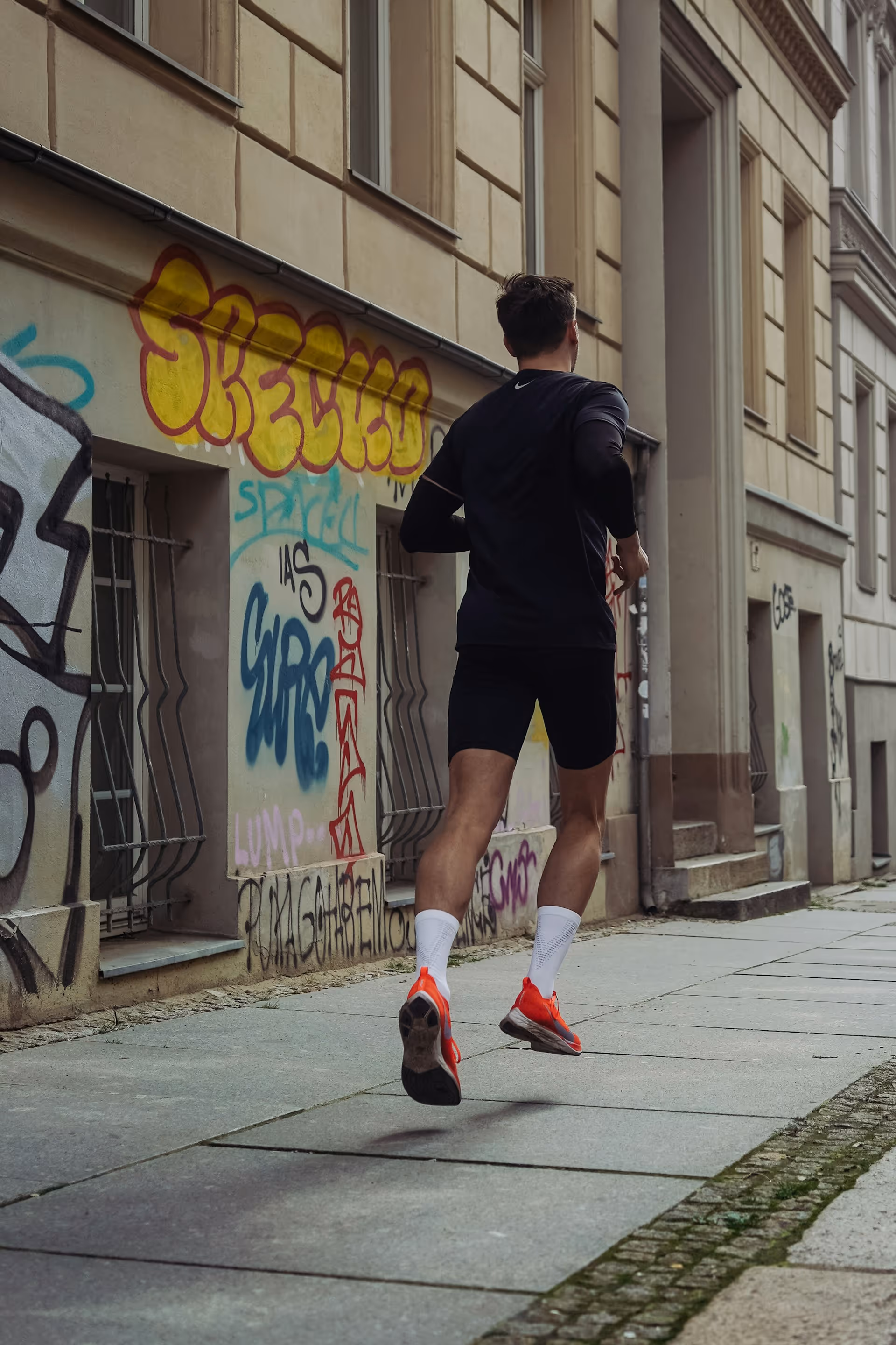 Man jogging on a paved urban street with graffiti-covered walls wearing black athletic clothing and bright orange running shoes.