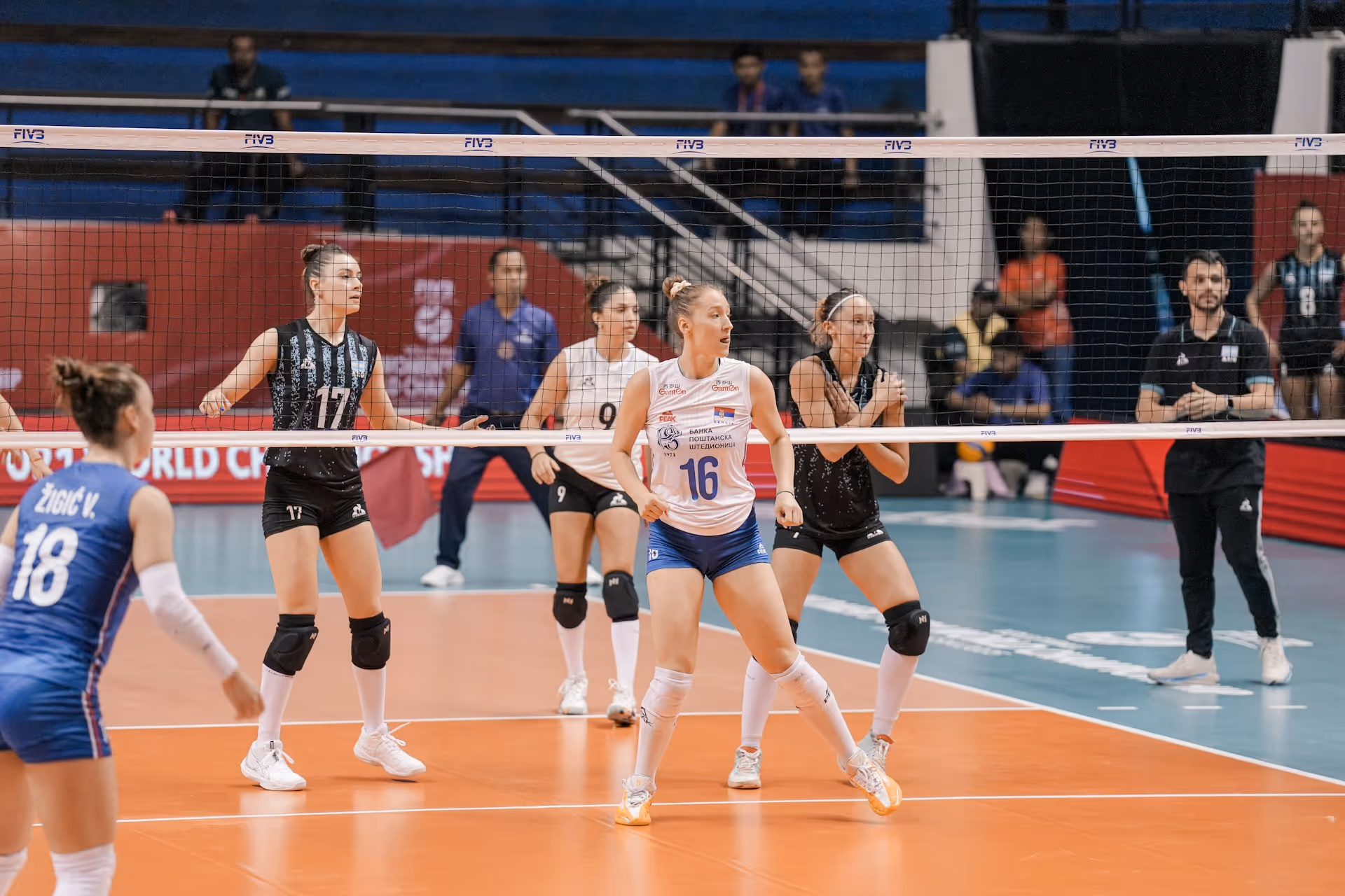Female volleyball players on court in action during a match, with coach and spectators in the background.