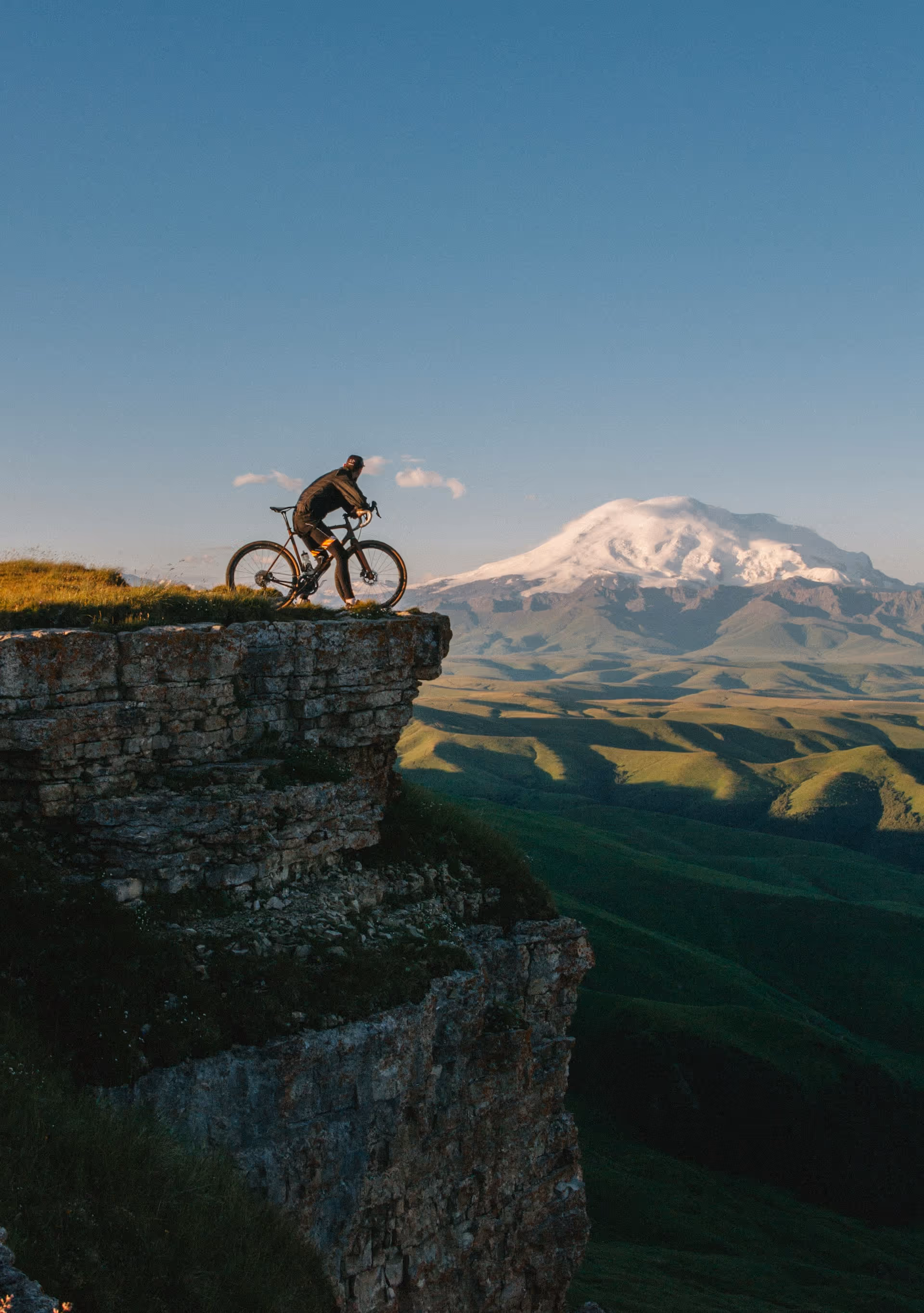 Person riding a bicycle on a grassy cliff edge overlooking green hills with a snow-capped mountain in the distance under a clear blue sky.