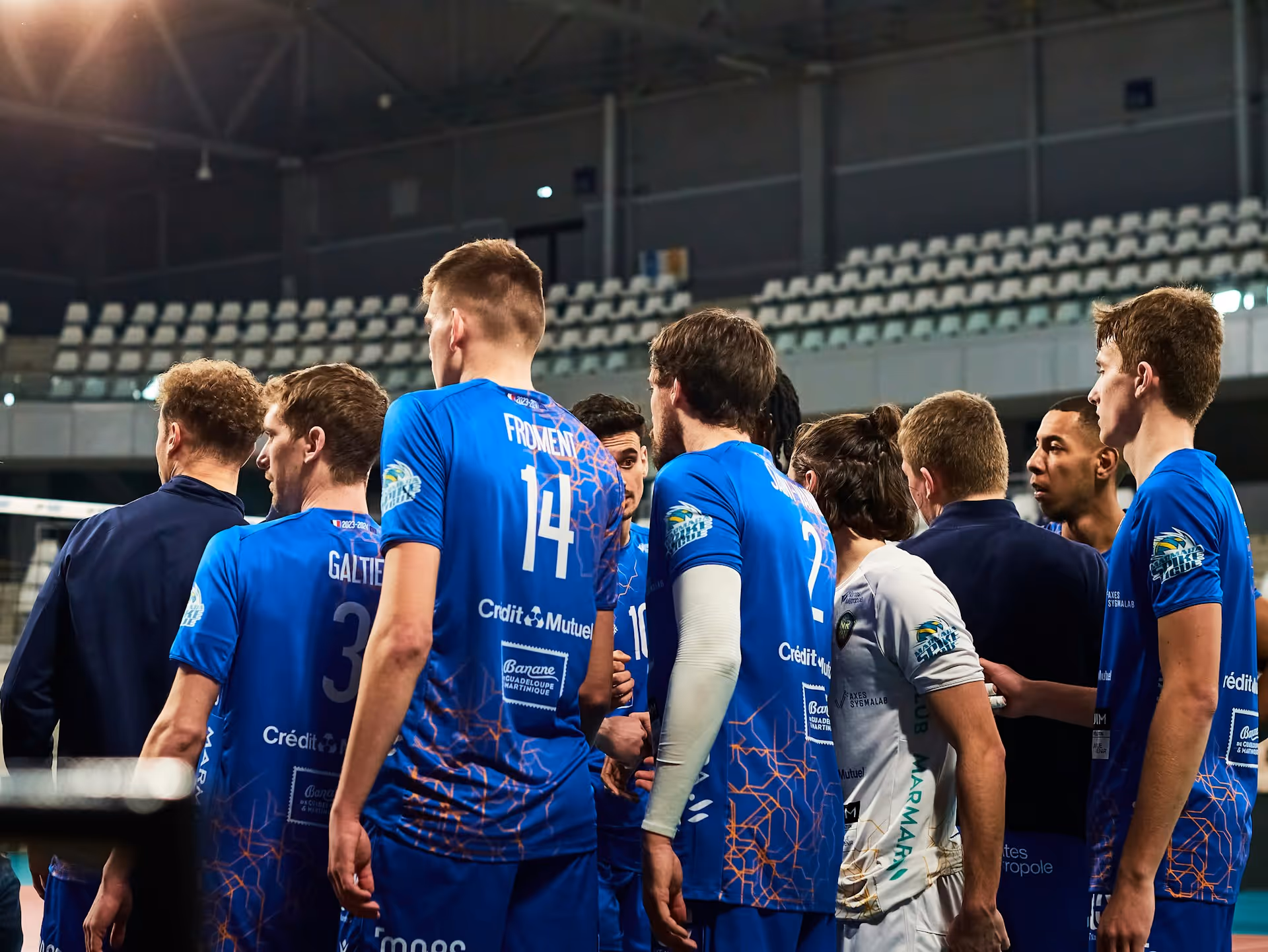 Male volleyball players in blue and white jerseys huddle together on an indoor court with empty stands in the background.