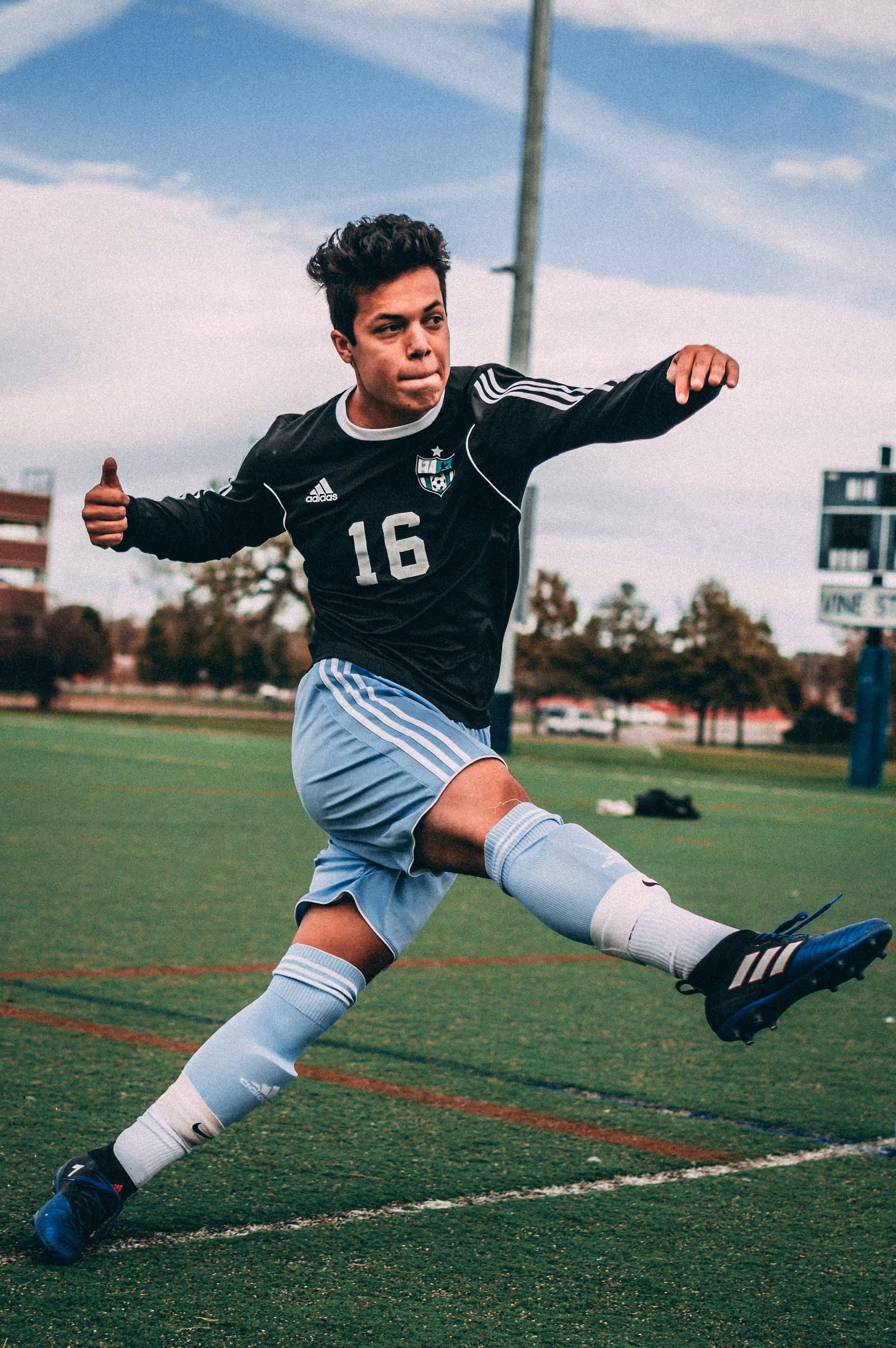 Soccer player wearing a black jersey with number 16 and light blue shorts kicking a ball on an outdoor field.