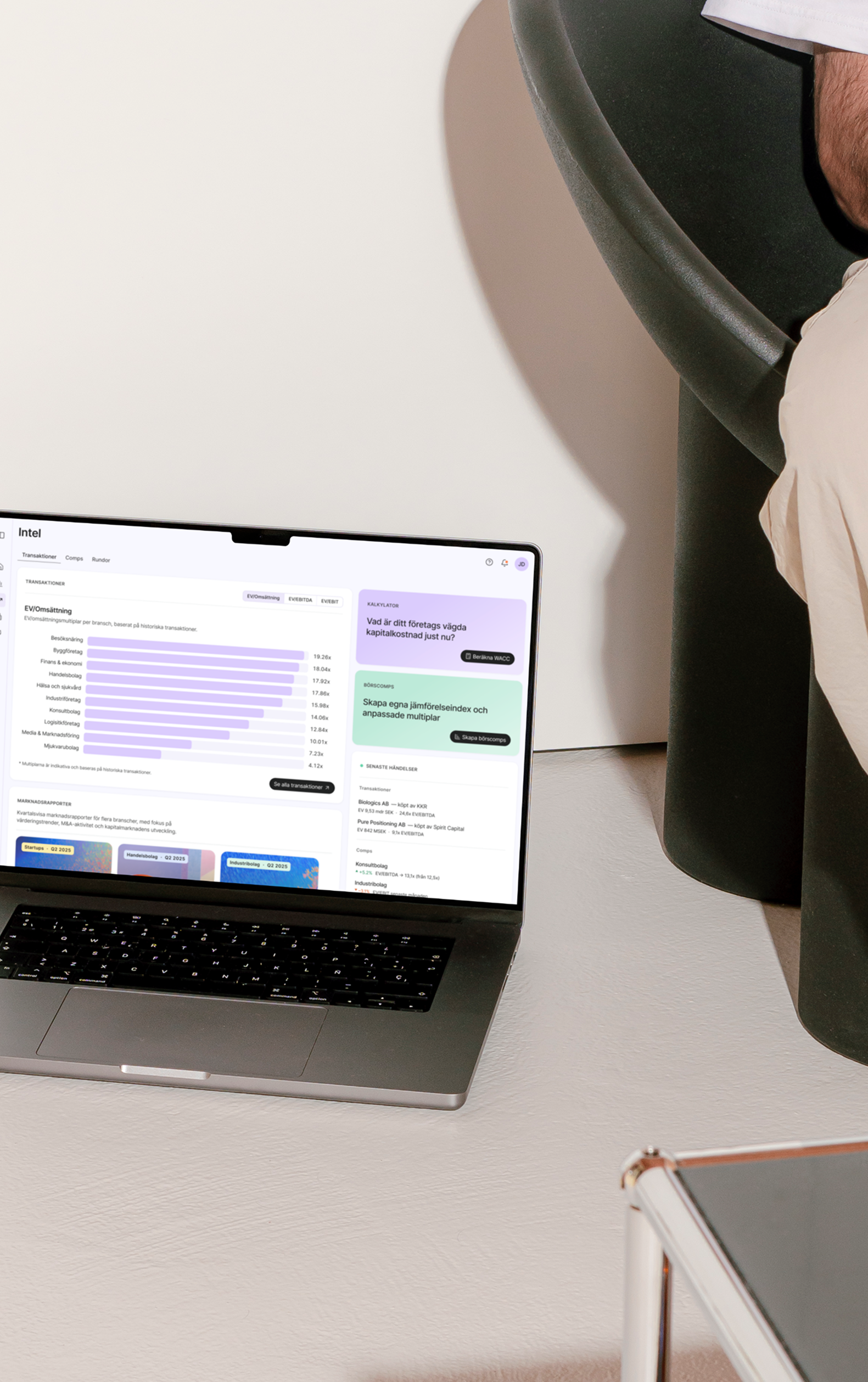 Open laptop on the floor displaying financial data and charts in Swedish, next to a green chair and a person's legs wearing beige pants.