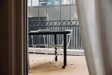 Outdoor patio with black metal table and two matching metal chairs on a beige carpet, partially viewed through a doorway with a curtain.