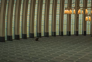 Interior of a spacious, curved mosque prayer hall with tall arched windows, green carpeted floor, and a single person sitting on the floor.