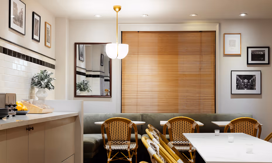 Cozy modern dining area with white tables, rattan chairs, a large window with wooden blinds, and framed black and white photos on the walls.