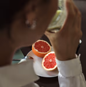 Close-up of a person holding a glass while looking at two halves of a grapefruit on a white plate.