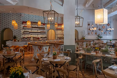 Cozy restaurant interior with wooden tables, chairs, hanging lanterns, and shelves lined with bottles.