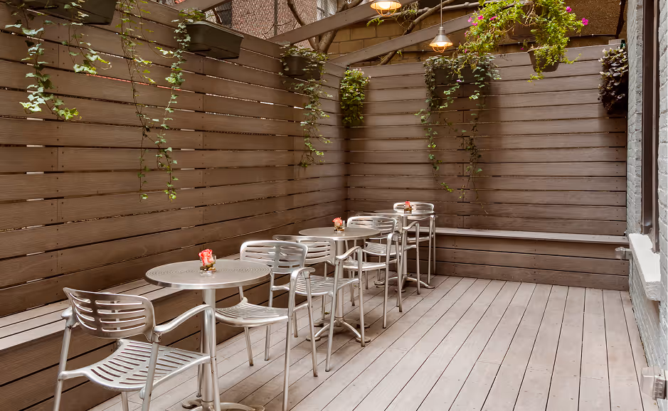Outdoor deck seating area with metal tables and chairs, wooden fence, hanging plants, and small floral centerpieces on tables.