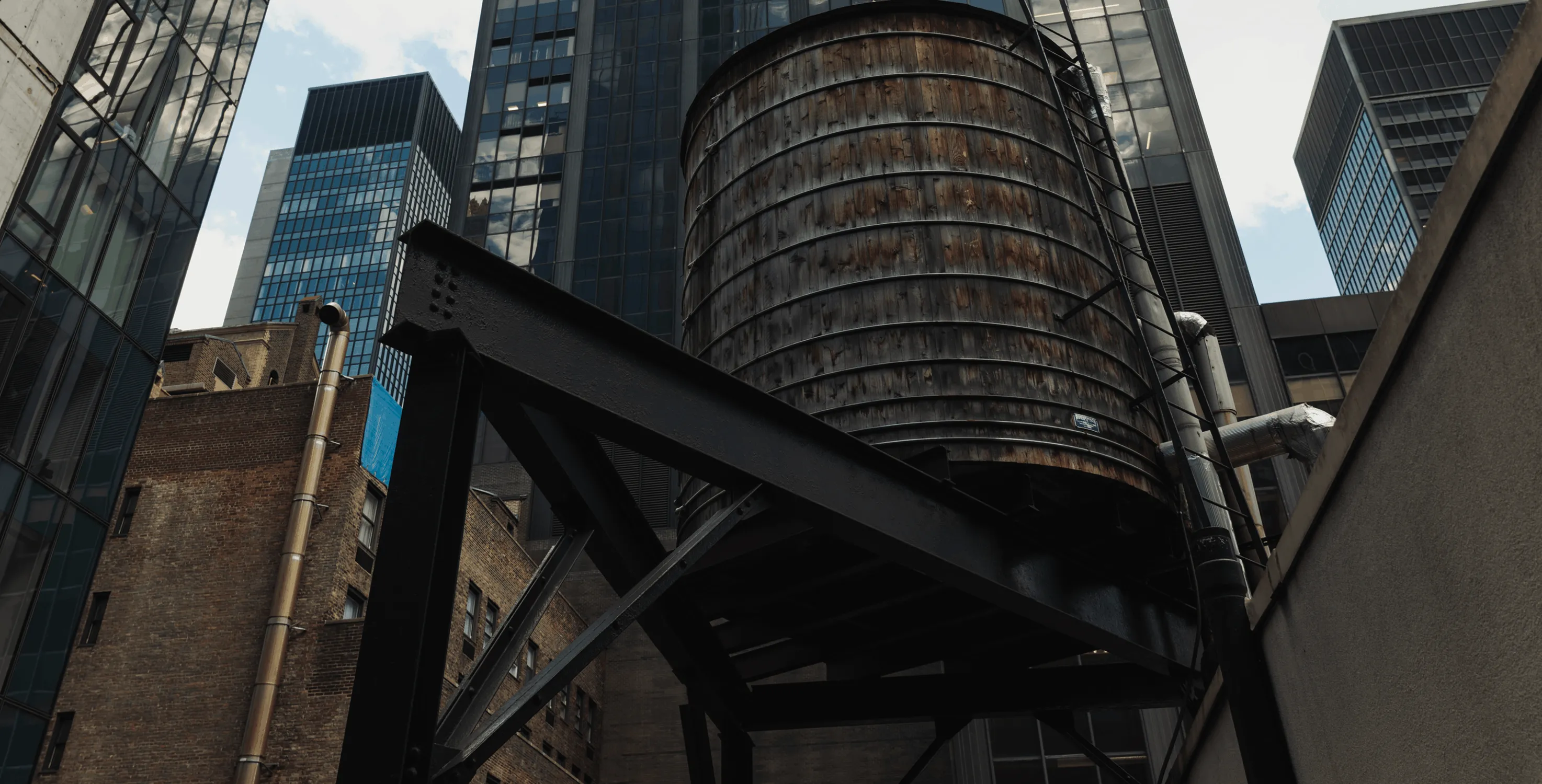Large cylindrical wooden water tank elevated on black metal supports amidst skyscrapers in a city.