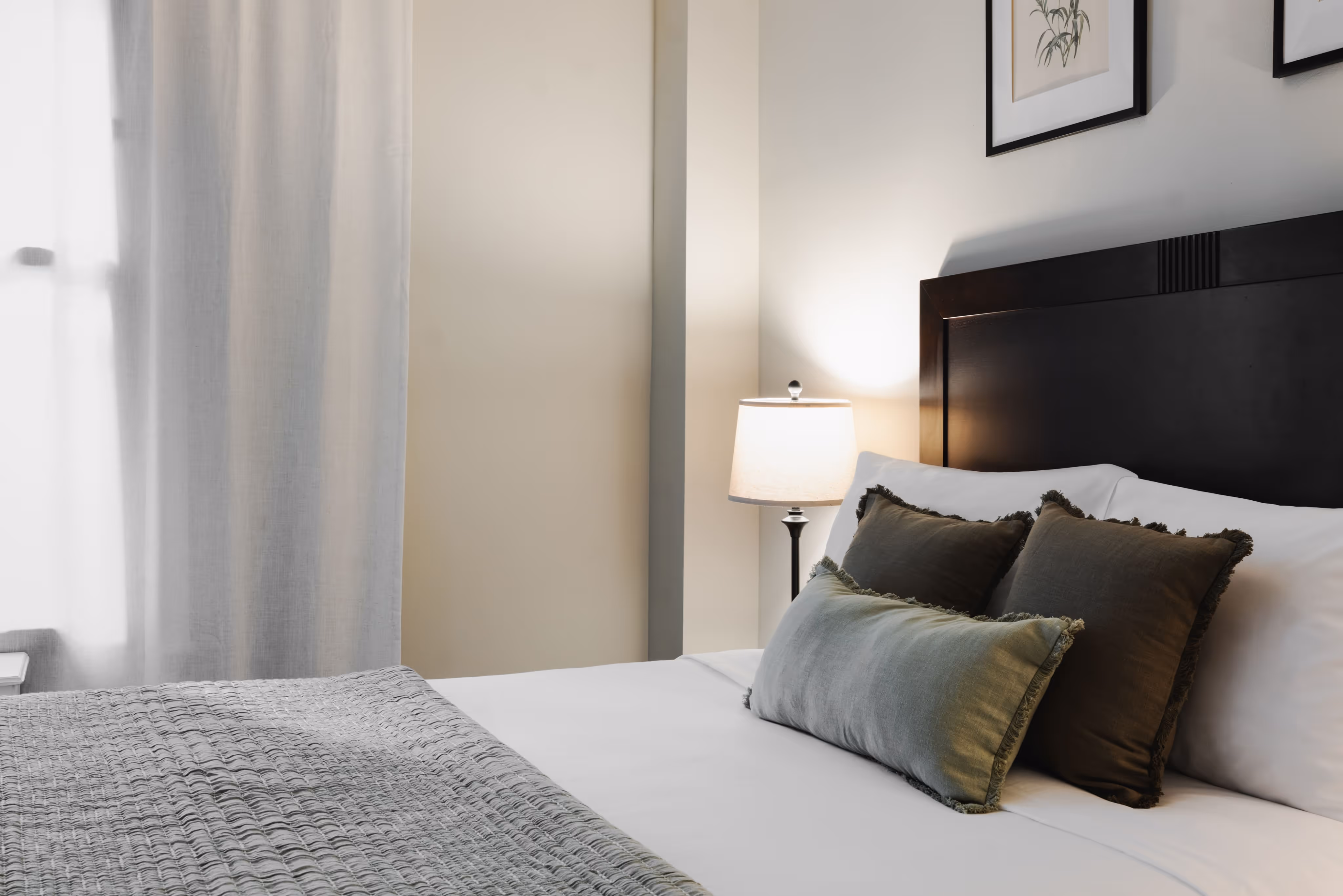 Side view of a neatly made bed with white sheets, three dark decorative pillows, a gray textured blanket, a lit bedside lamp, and framed botanical prints on a light-colored wall.