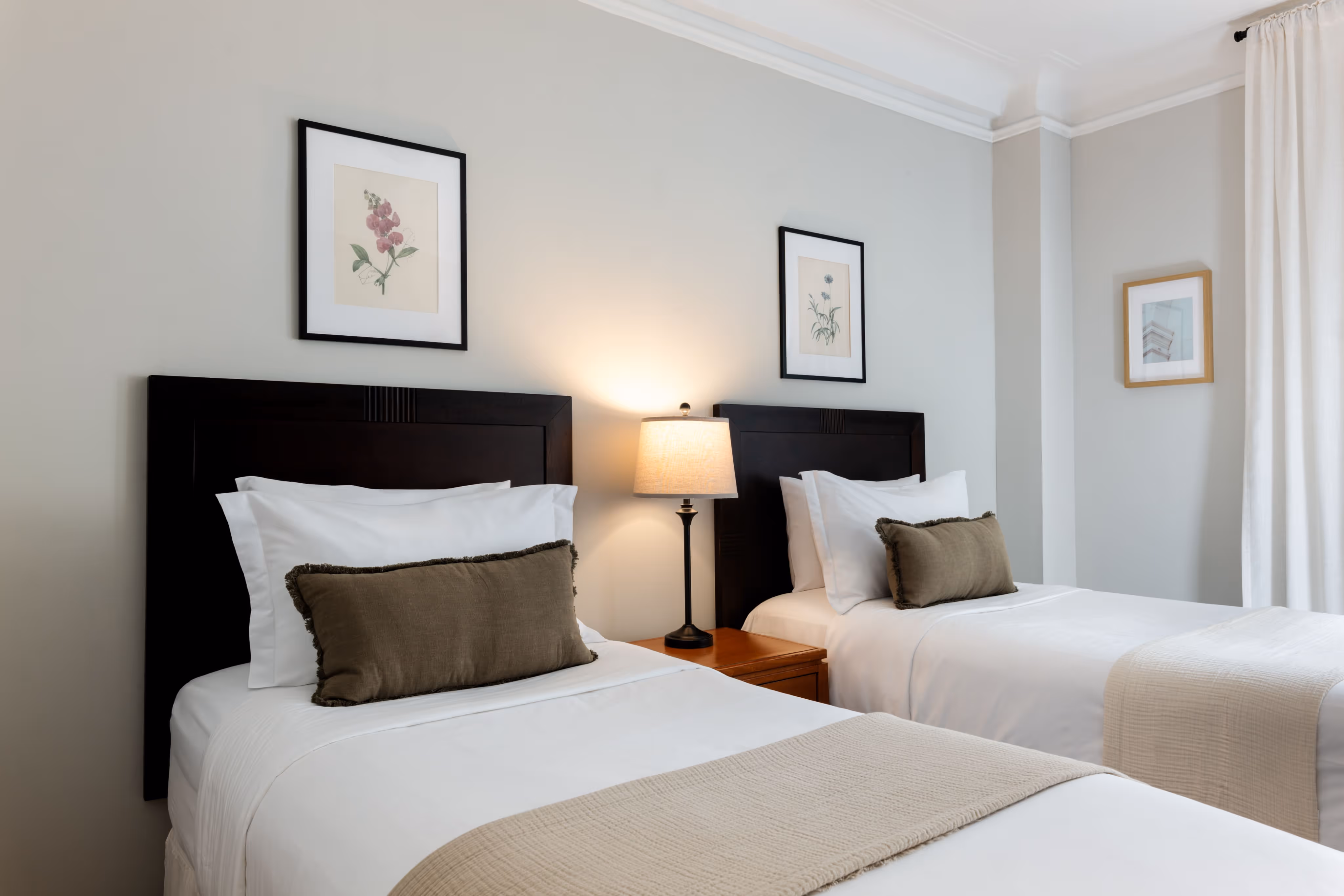 Two neatly made twin beds with dark wood headboards, beige throw pillows, and a lit bedside lamp between them.