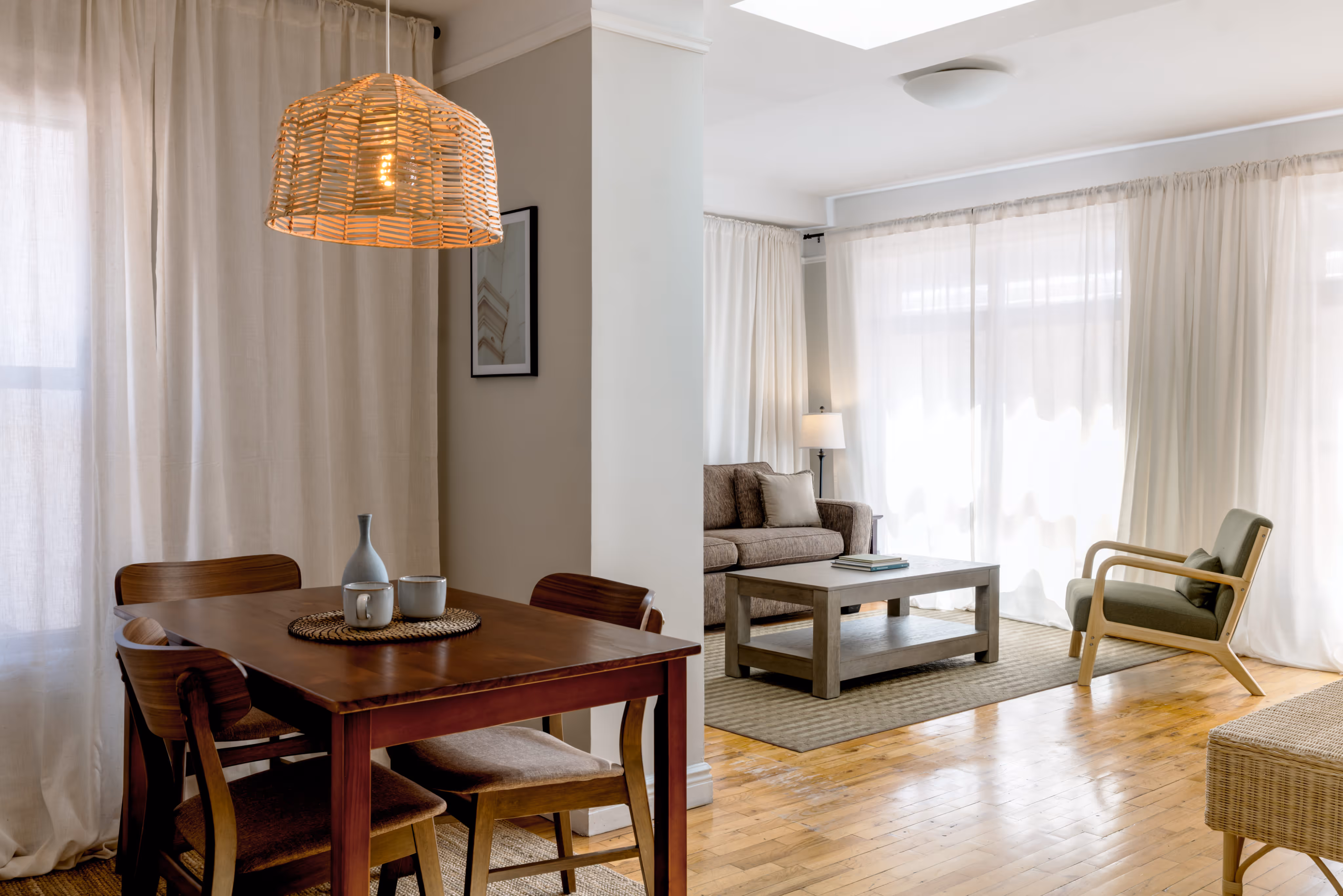 Bright living and dining area with wooden dining table, four chairs, pendant light, sofa, armchair, coffee table, and large windows with sheer curtains.