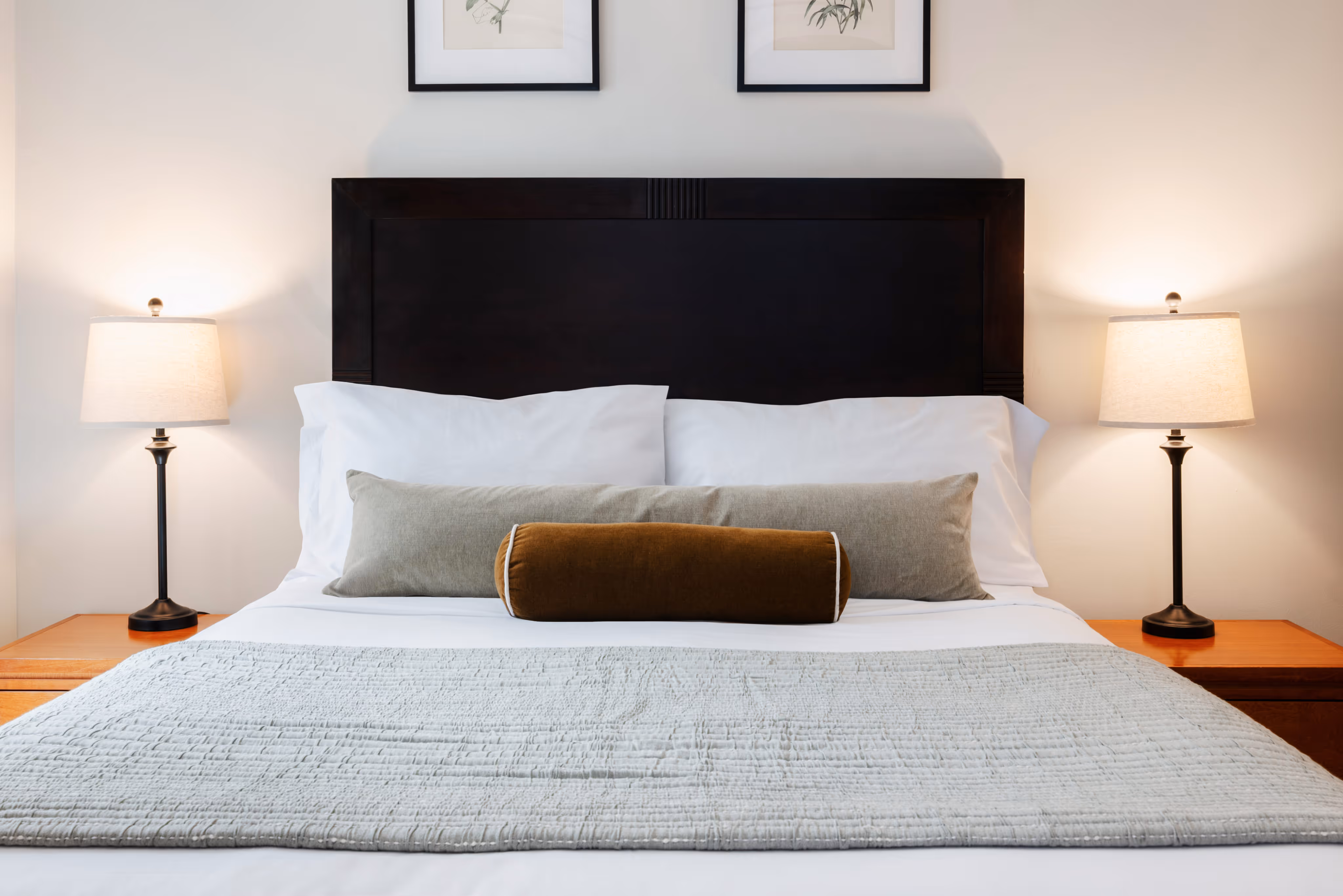 Neatly made bed with a dark wooden headboard, white pillows, two decorative cushions, and two lit bedside lamps on wooden tables.