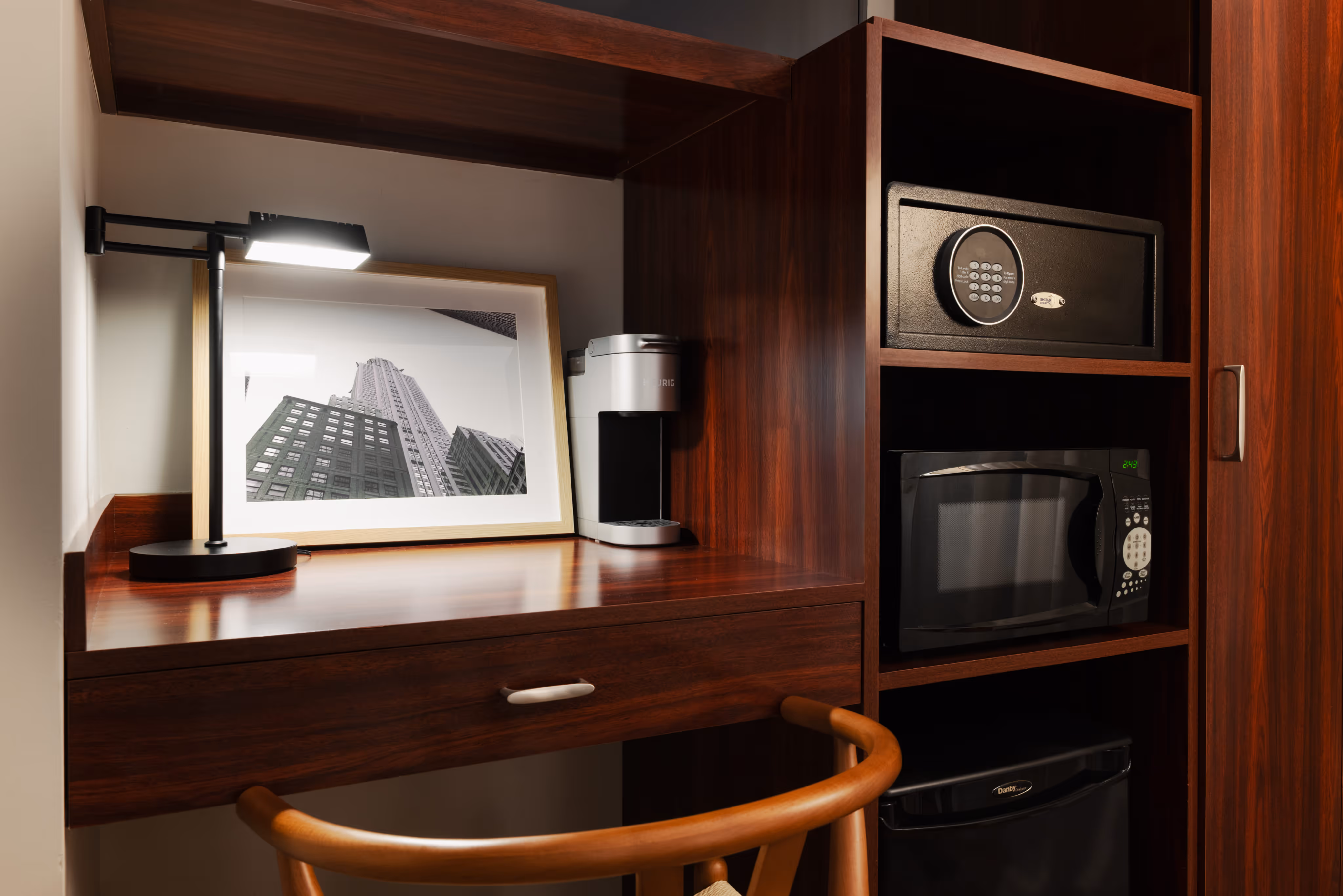Wooden desk with a modern desk lamp, framed black and white photo of skyscrapers, coffee maker, adjacent cabinet with electronic safe, microwave, and mini fridge.