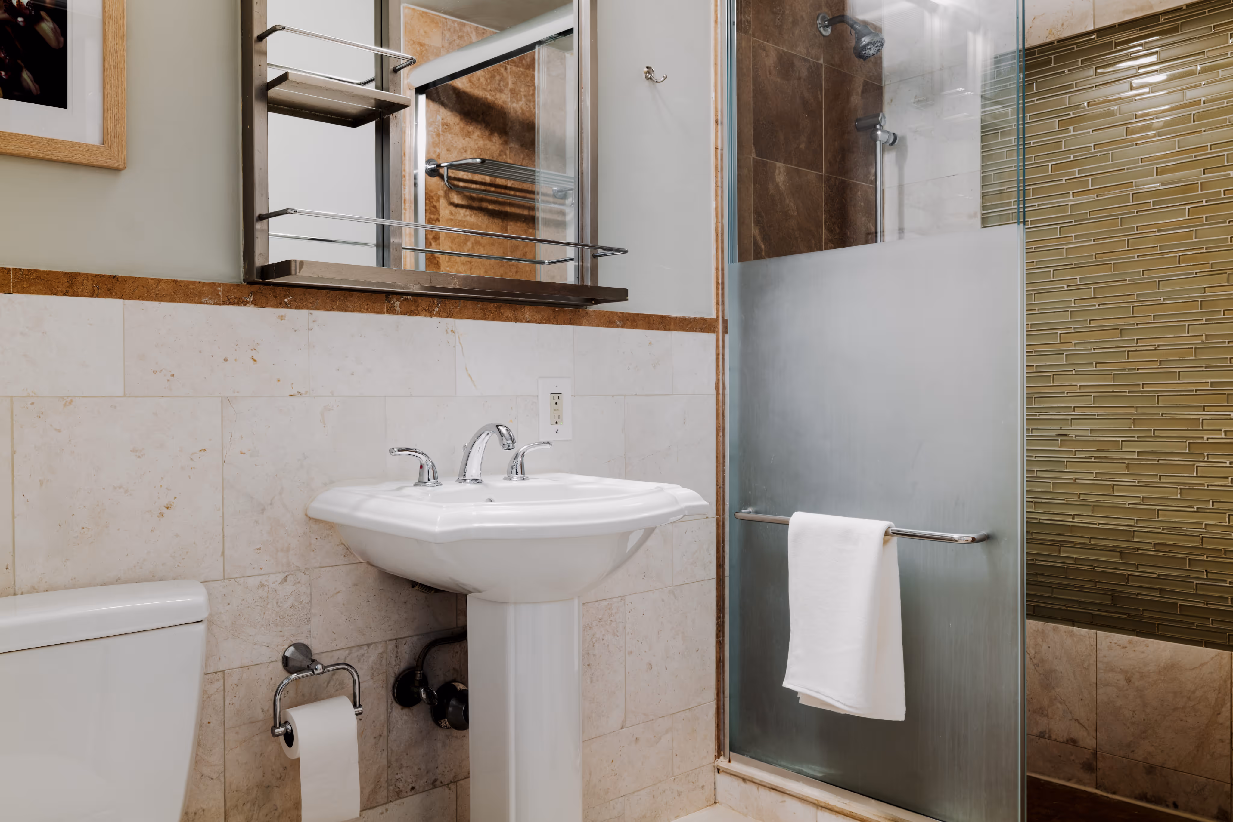 Modern bathroom with white pedestal sink, toilet, frosted glass shower door with towel, and beige tiled walls.
