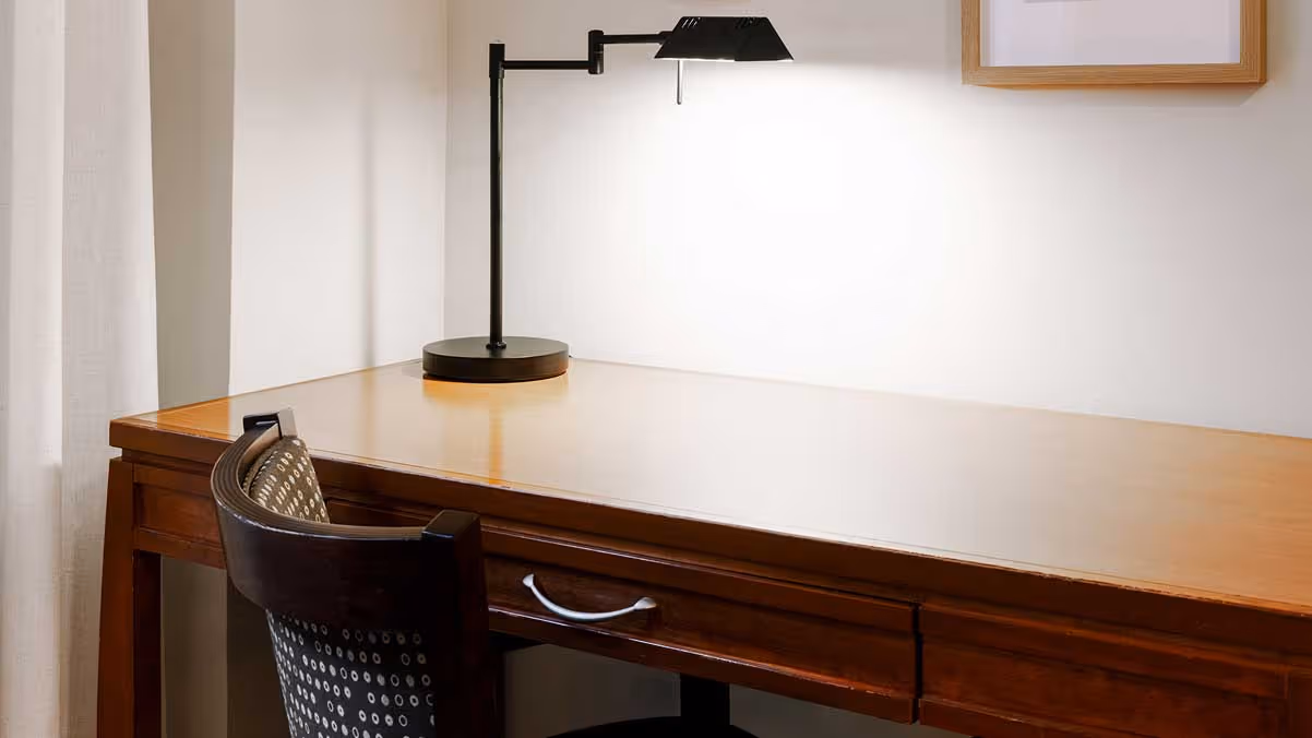 Wooden desk with a black patterned chair, a black desk lamp, a round wooden-framed mirror, and a framed botanical print on a beige wall.