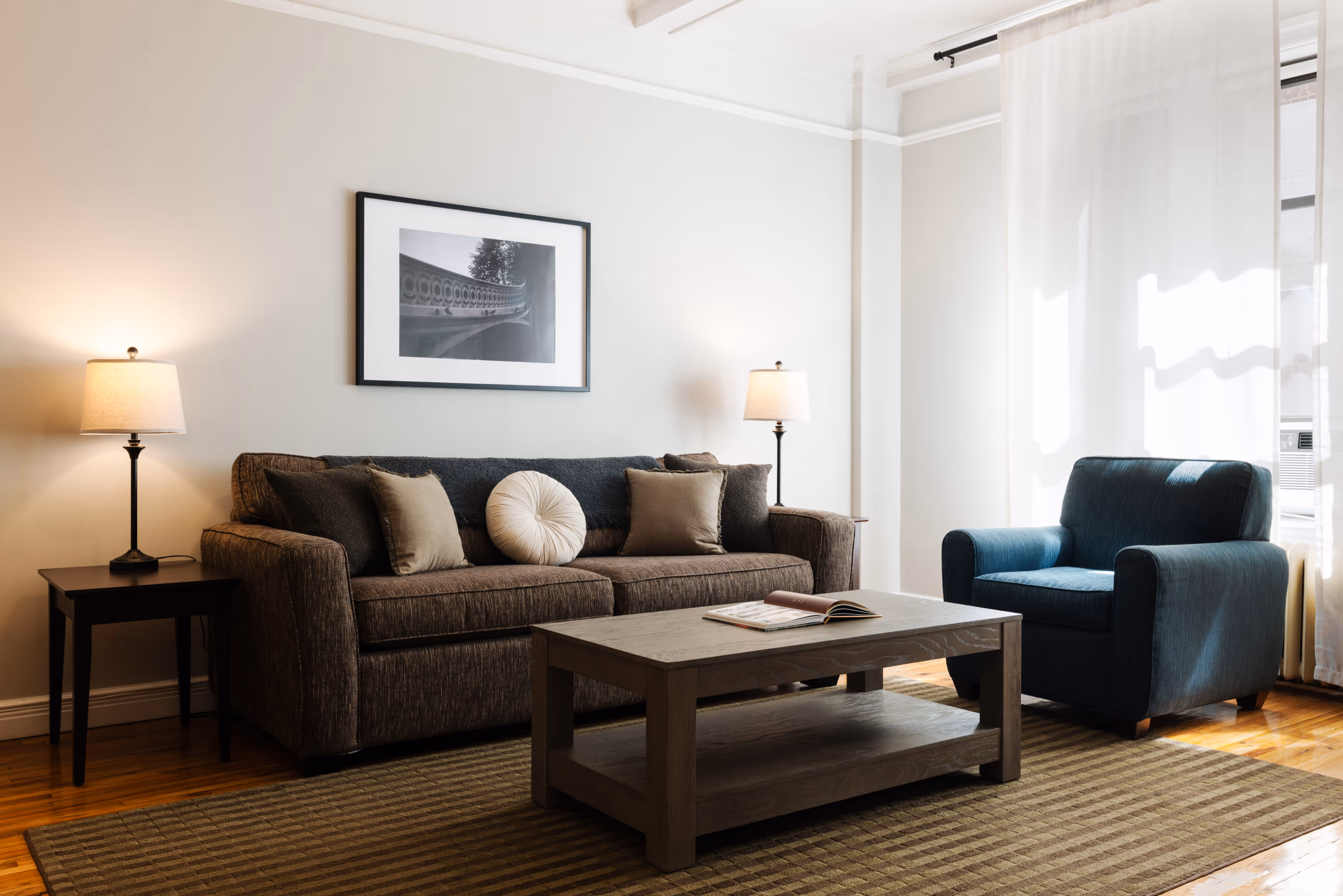 Modern living room with a brown sofa adorned with cushions, a blue armchair, wooden coffee table with an open book, two side lamps, and a framed black-and-white photo on the wall.
