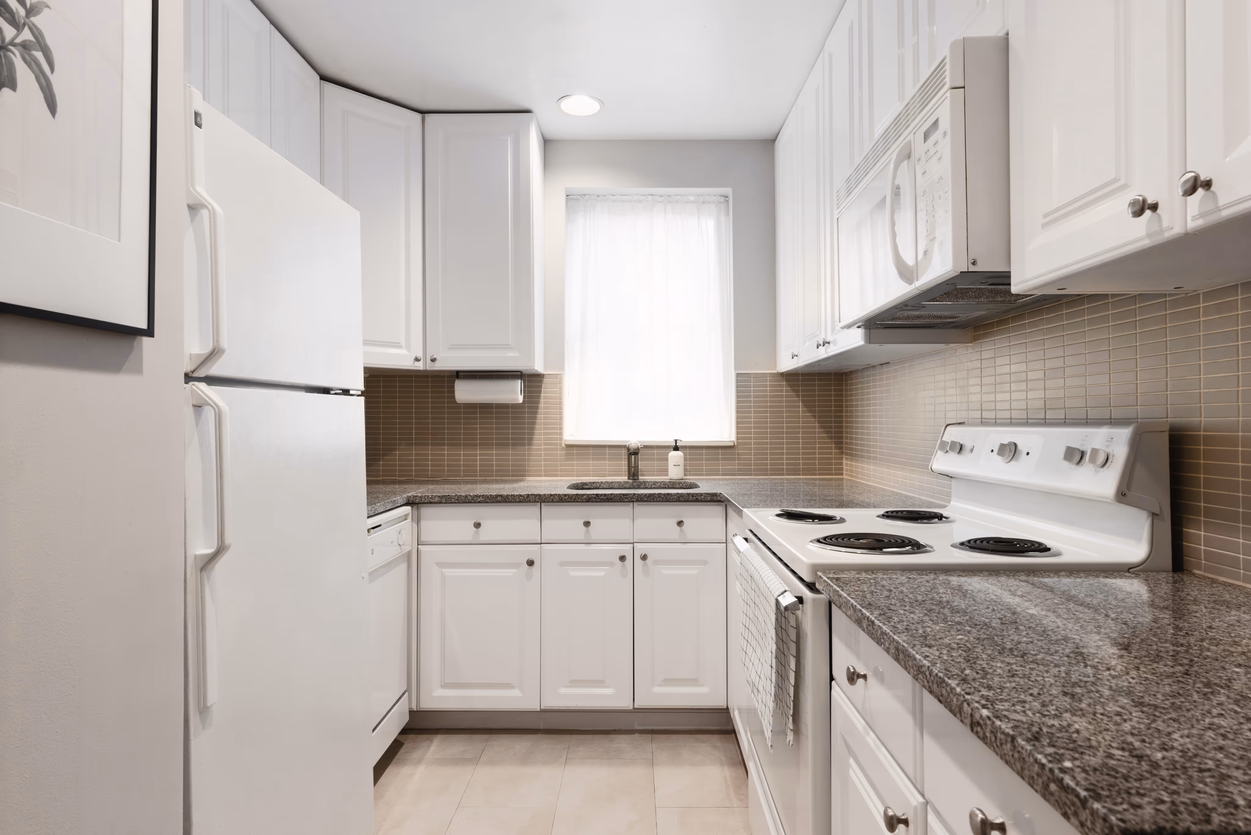 Compact U-shaped kitchen with white cabinets, granite countertops, white appliances, and a window with sheer curtain above the sink.