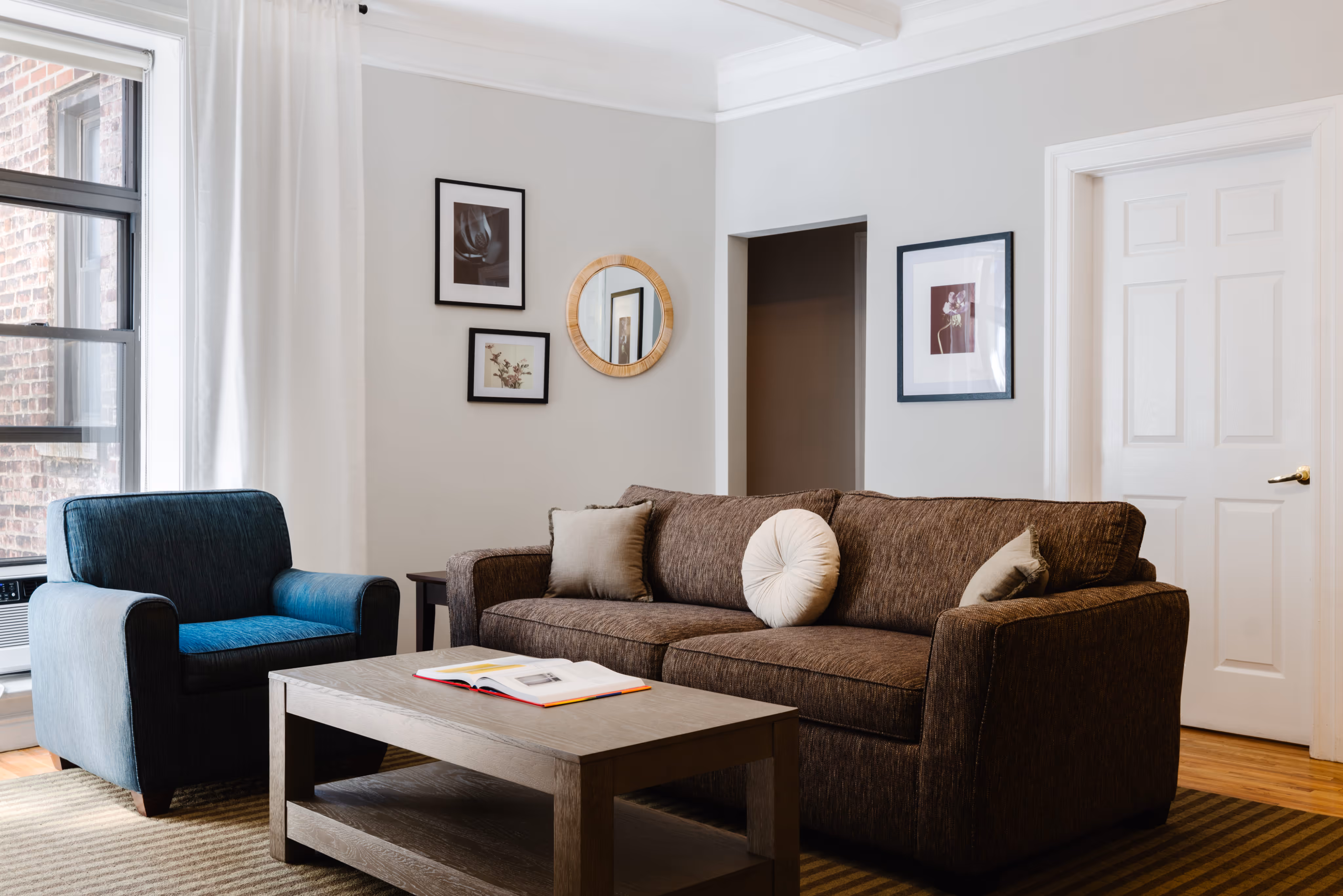 Living room with a brown sofa, blue armchair, wooden coffee table with an open book, and framed artwork on light-colored walls.