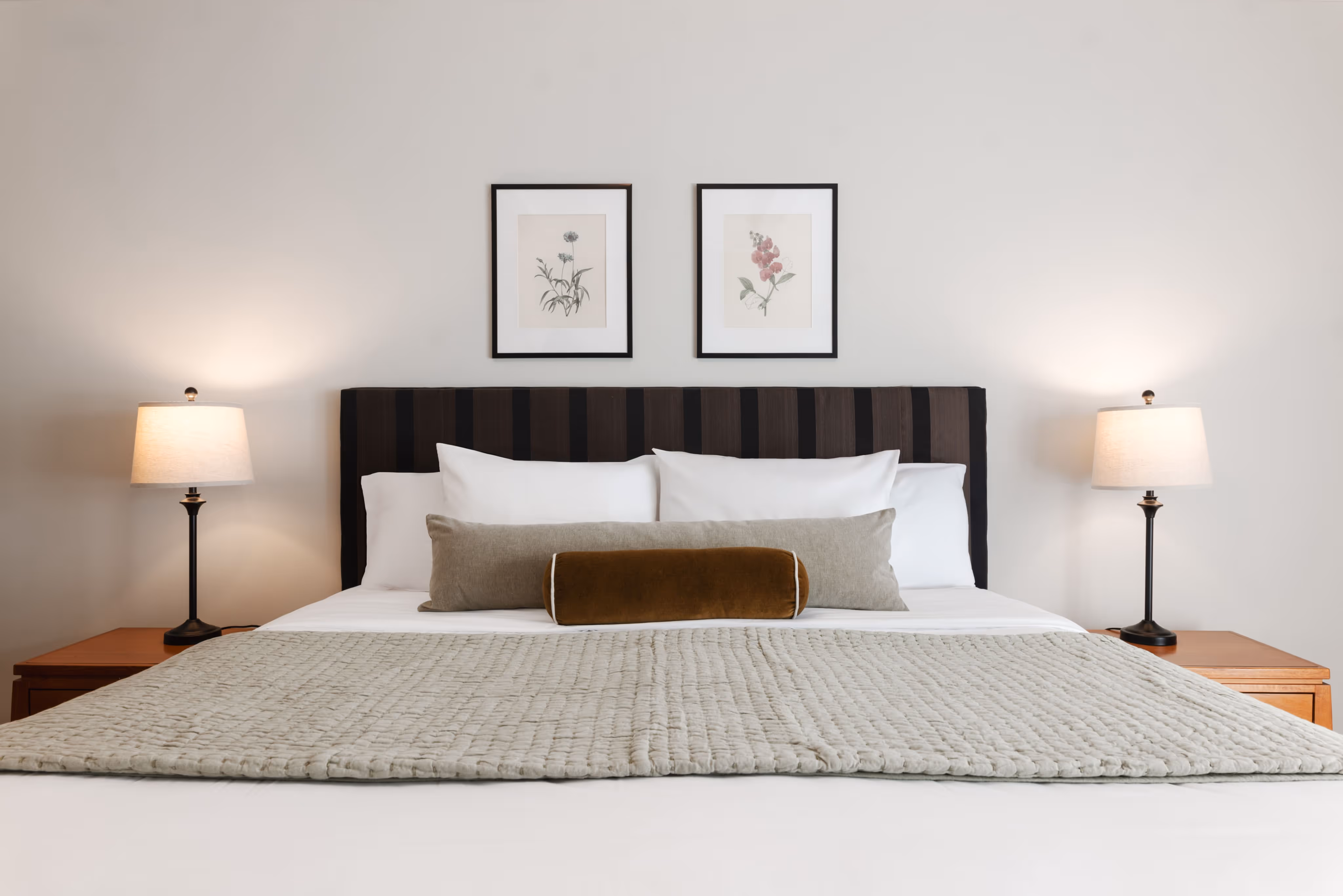Modern bed with white sheets, multiple pillows, and a textured beige throw, flanked by two wooden nightstands with lit lamps and floral artwork above the headboard.