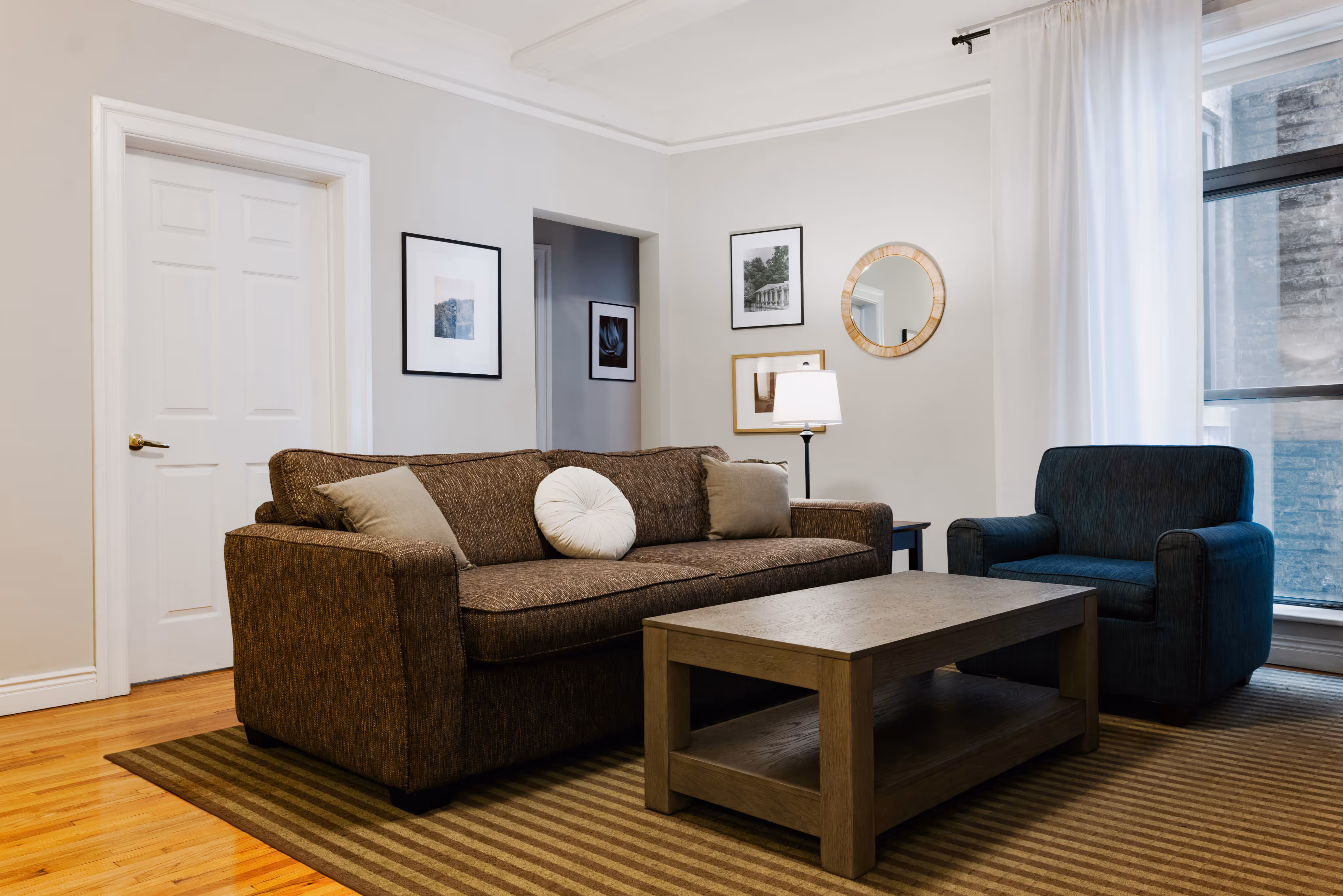 Living room with brown sofa, blue armchair, wooden coffee table, striped rug, framed pictures, and large window with white curtains.