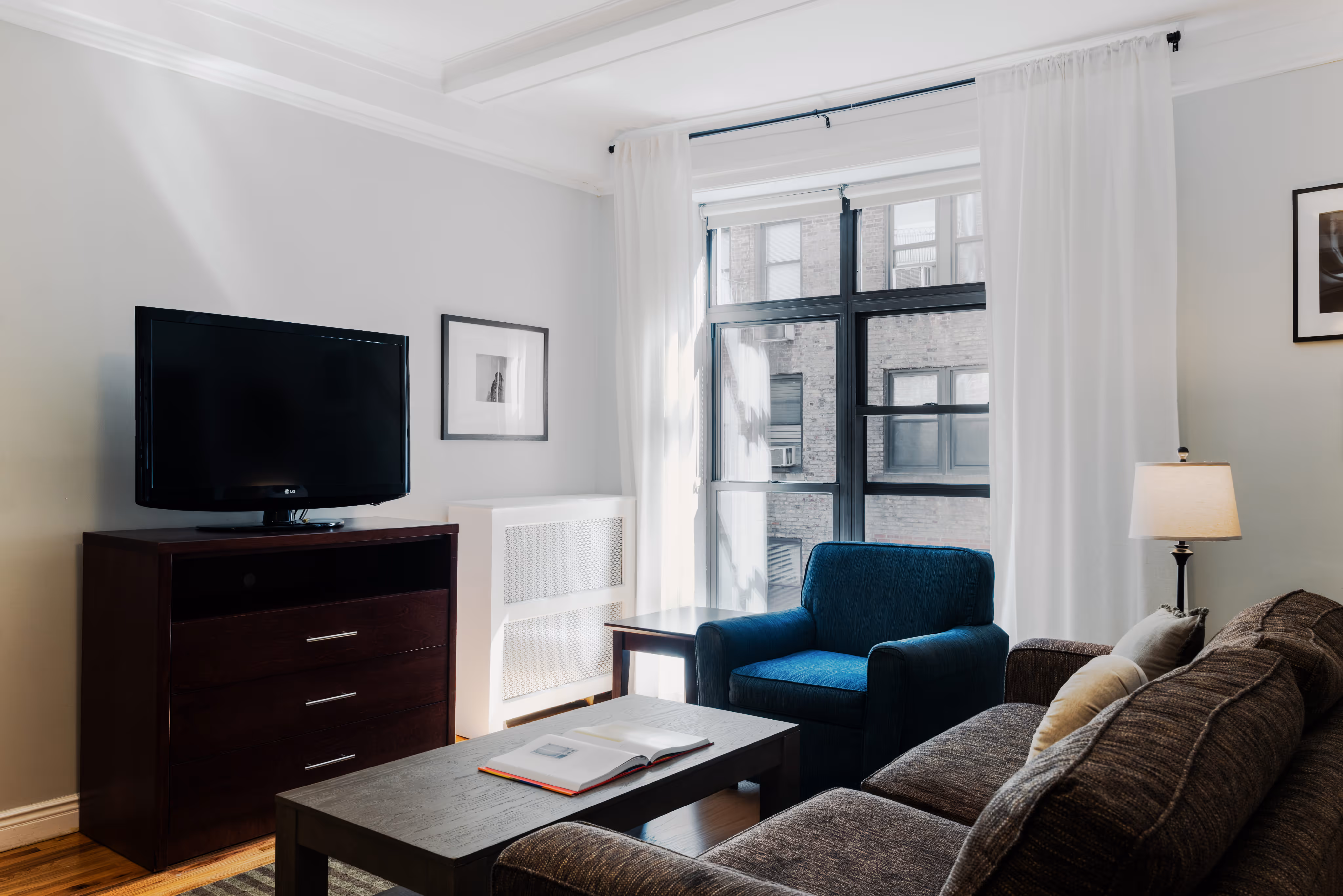 Living room with a dark brown sofa, blue armchair, coffee table with an open book, TV on a stand, and large window with white curtains.