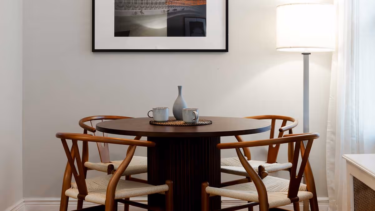 Minimalist dining area with round dark wooden table, four wooden chairs with woven seats, pendant light, floor lamp, and framed artwork on white wall.