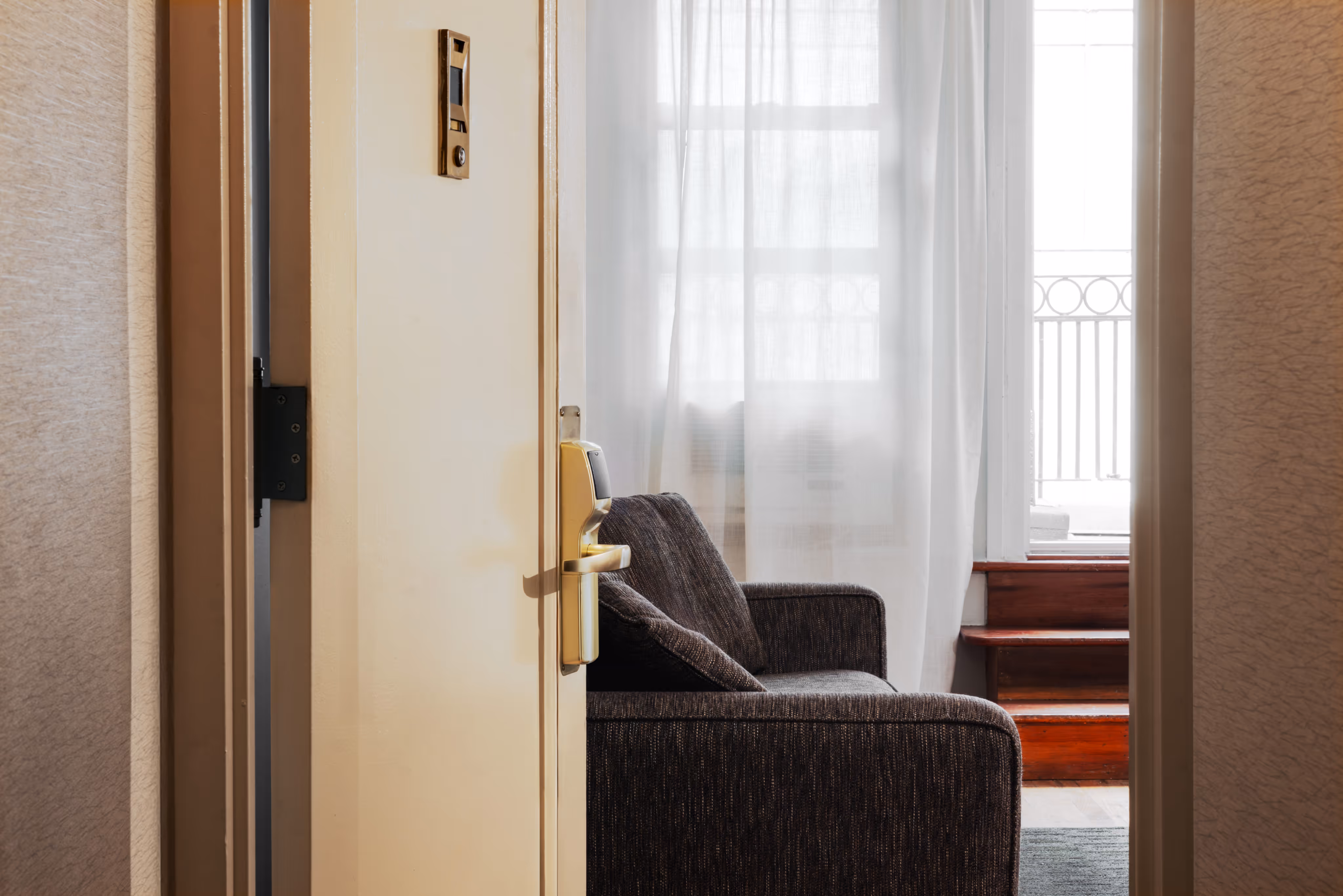 View through an open door showing a dark gray armchair, sheer white curtains, and wooden stairs leading to a balcony.
