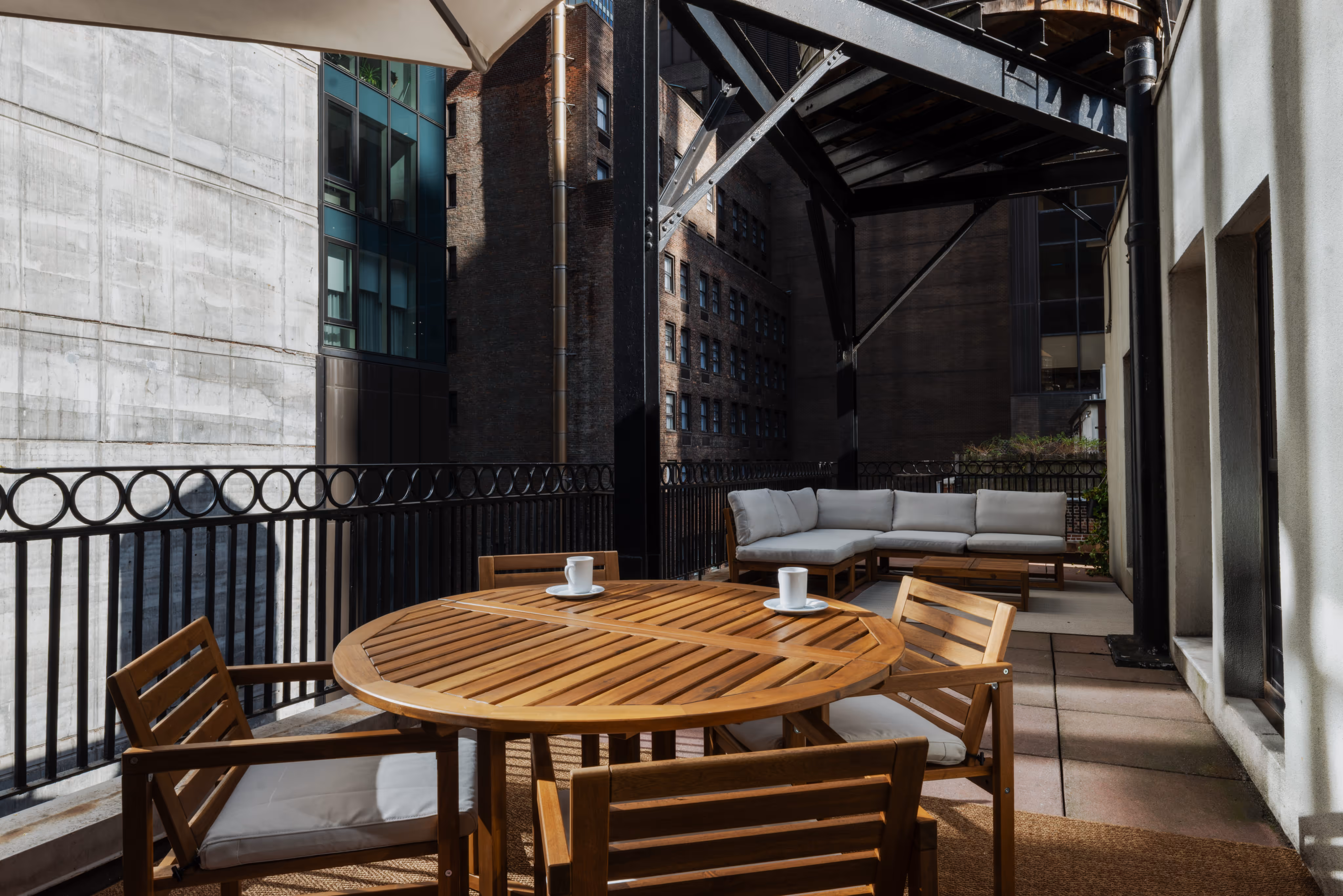 Outdoor balcony seating area with a wooden round table, four chairs with cushions, two coffee cups, and a cushioned corner sofa under an umbrella and metal framework.